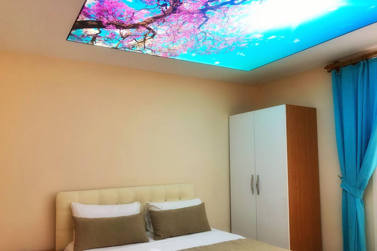 A hotel room with a bed, a wardrobe, a nightstand, and a ceiling with a tree and sky view.