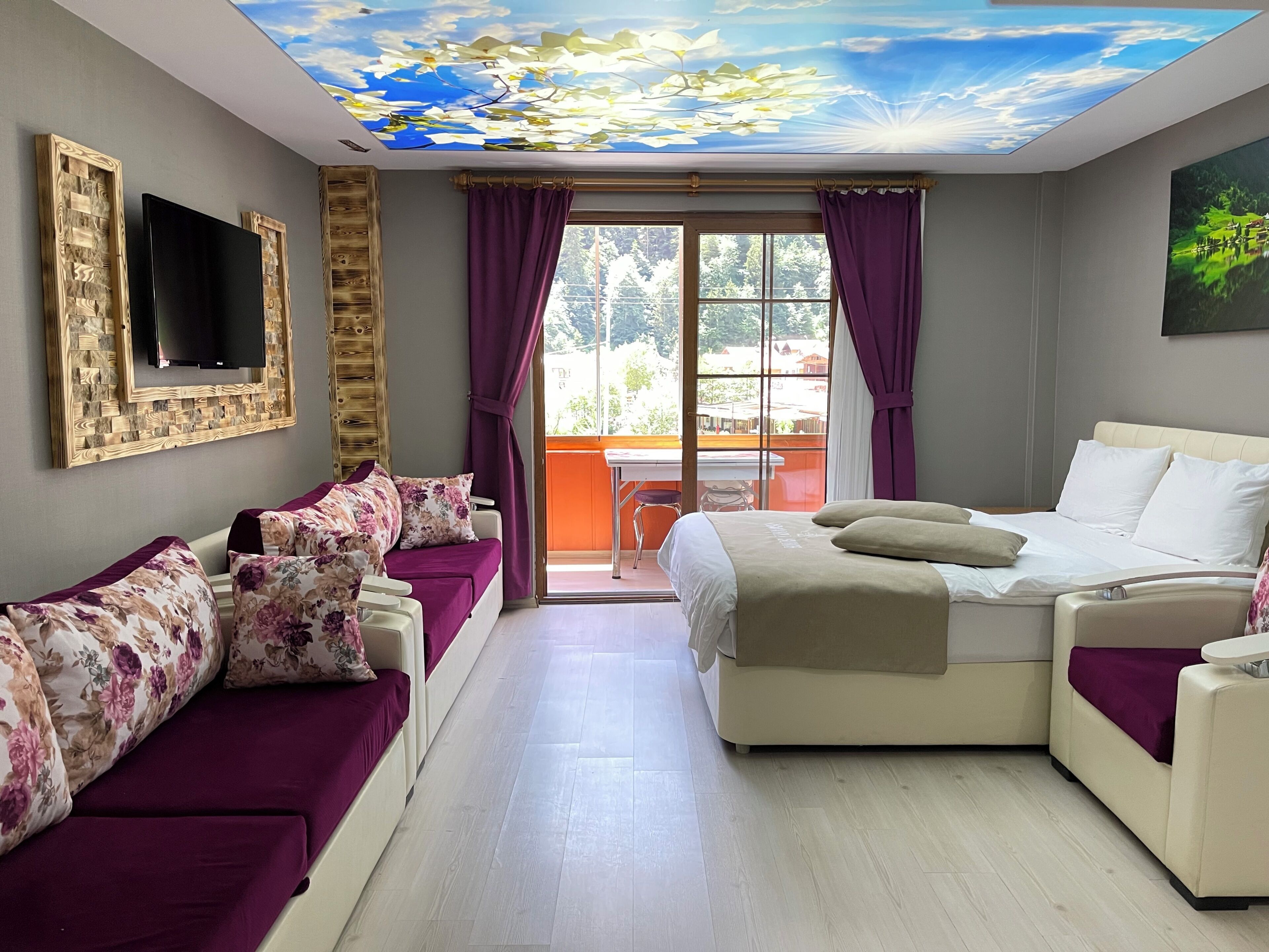A hotel room with a bed, sofa, TV, ceiling artwork, and a balcony view.