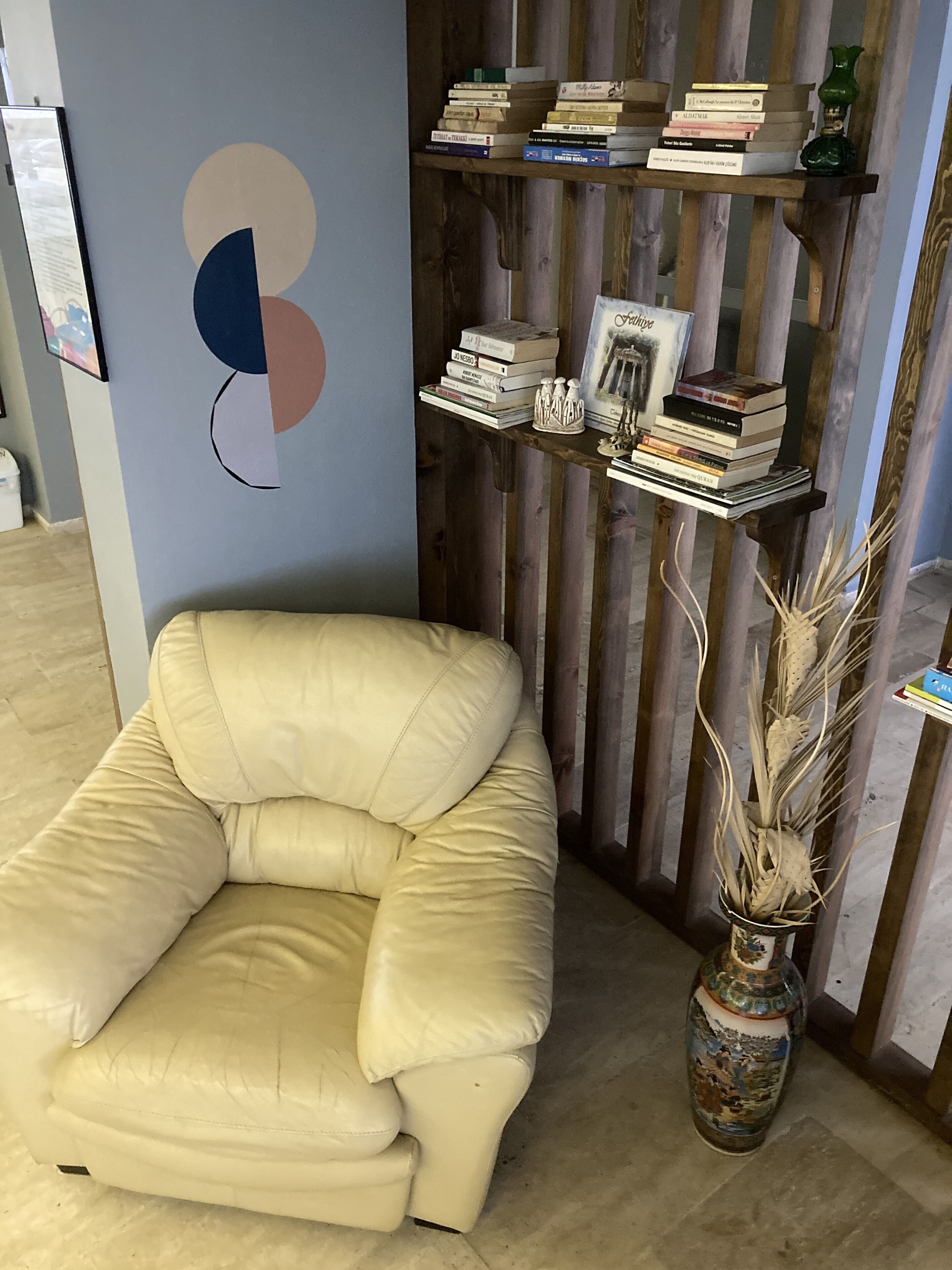 Ein beiger Sessel, ein Holzregal mit Büchern und eine dekorative Vase mit Trockenpflanzen.