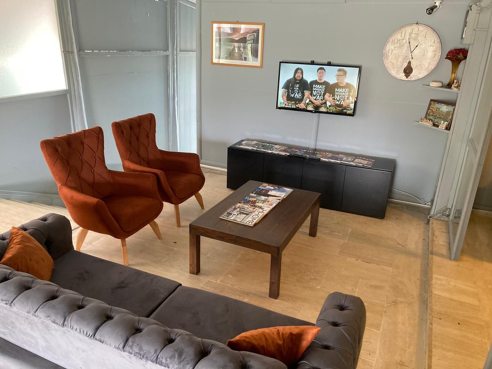 Una sala de estar moderna con un sofá gris, dos sillones naranjas, una mesa de centro de madera, un televisor montado en la pared y una estantería con revistas.