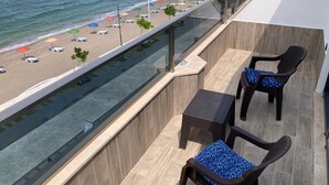 Un balcón con dos sillas de mimbre y una mesita, con vista a una playa con sombrillas y tumbonas.