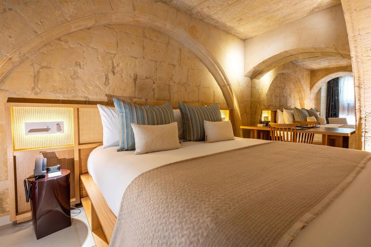 A room with a large bed, a wooden desk, and a stone wall.