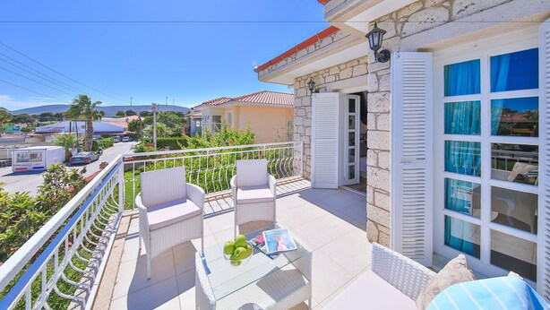A balcony with white wicker furniture, a glass table with green apples and a magazine, and a stone building with a red roof in the background. - Alacati Sorf Hotel (Cesme)