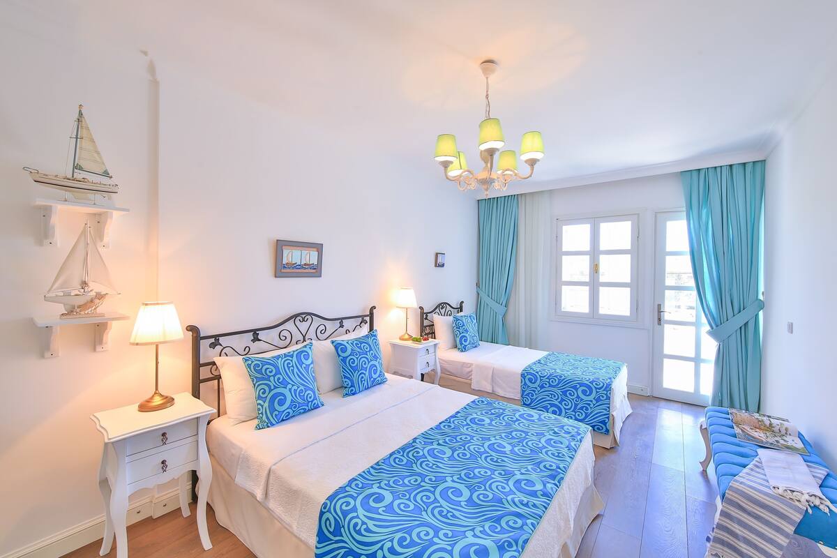 A hotel room with two beds, a chandelier, a window with curtains, and a small shelf with a model ship.