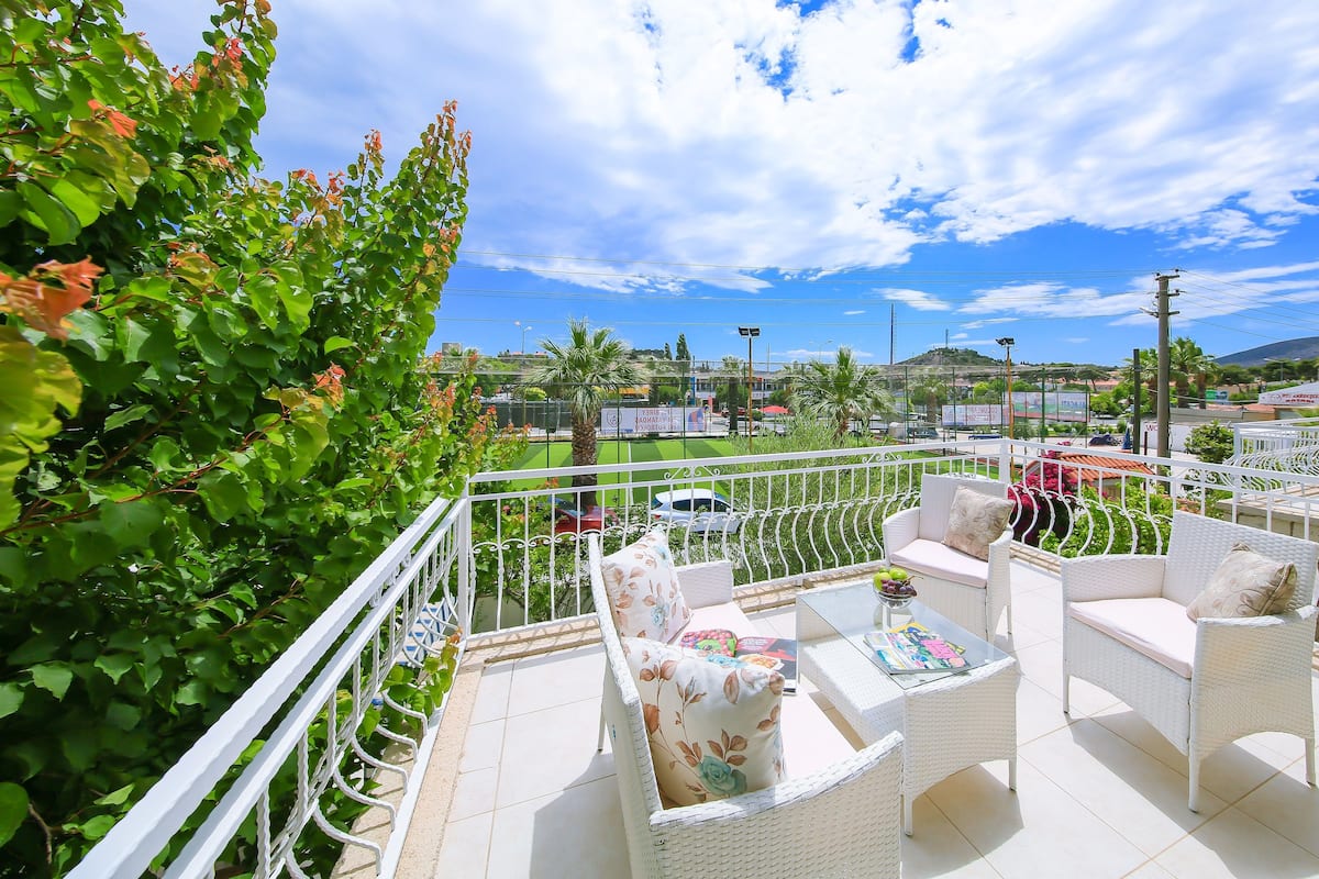 A balcony with white wicker furniture, a table with a bowl of fruit, and a view of a cityscape with buildings and palm trees.