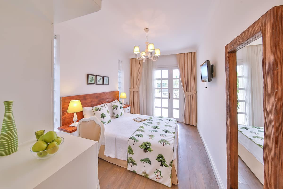 A bedroom with a wooden headboard, a bedside table with a lamp, a bed with a tree-patterned bedspread, a large mirror, and a television on the wall.