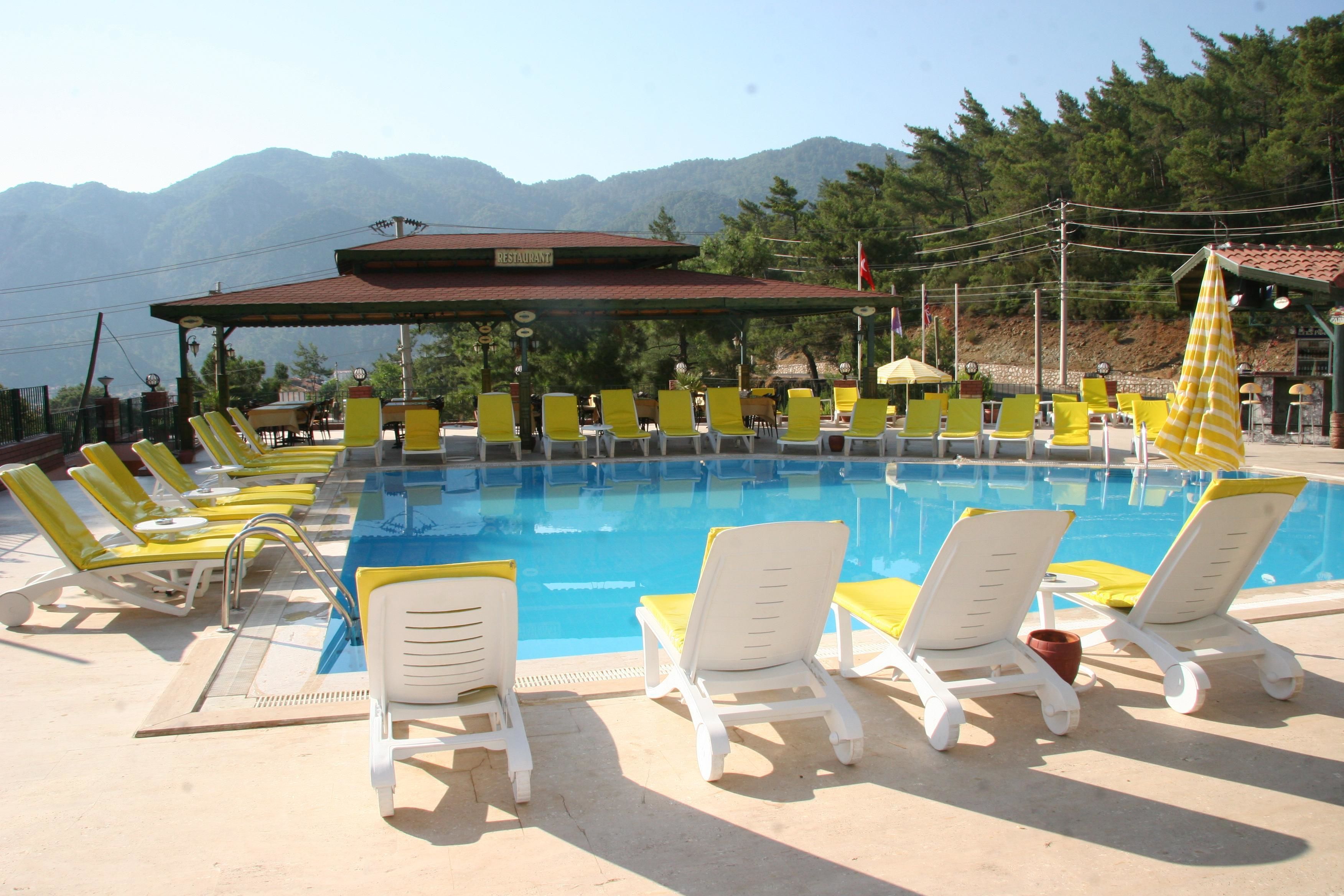 Zona de piscina con sillones reclinables amarillos y blancos, un restaurante y montañas al fondo.