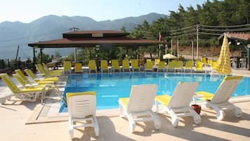 Zona de piscina con sillones reclinables amarillos y blancos, un restaurante y montañas al fondo.
