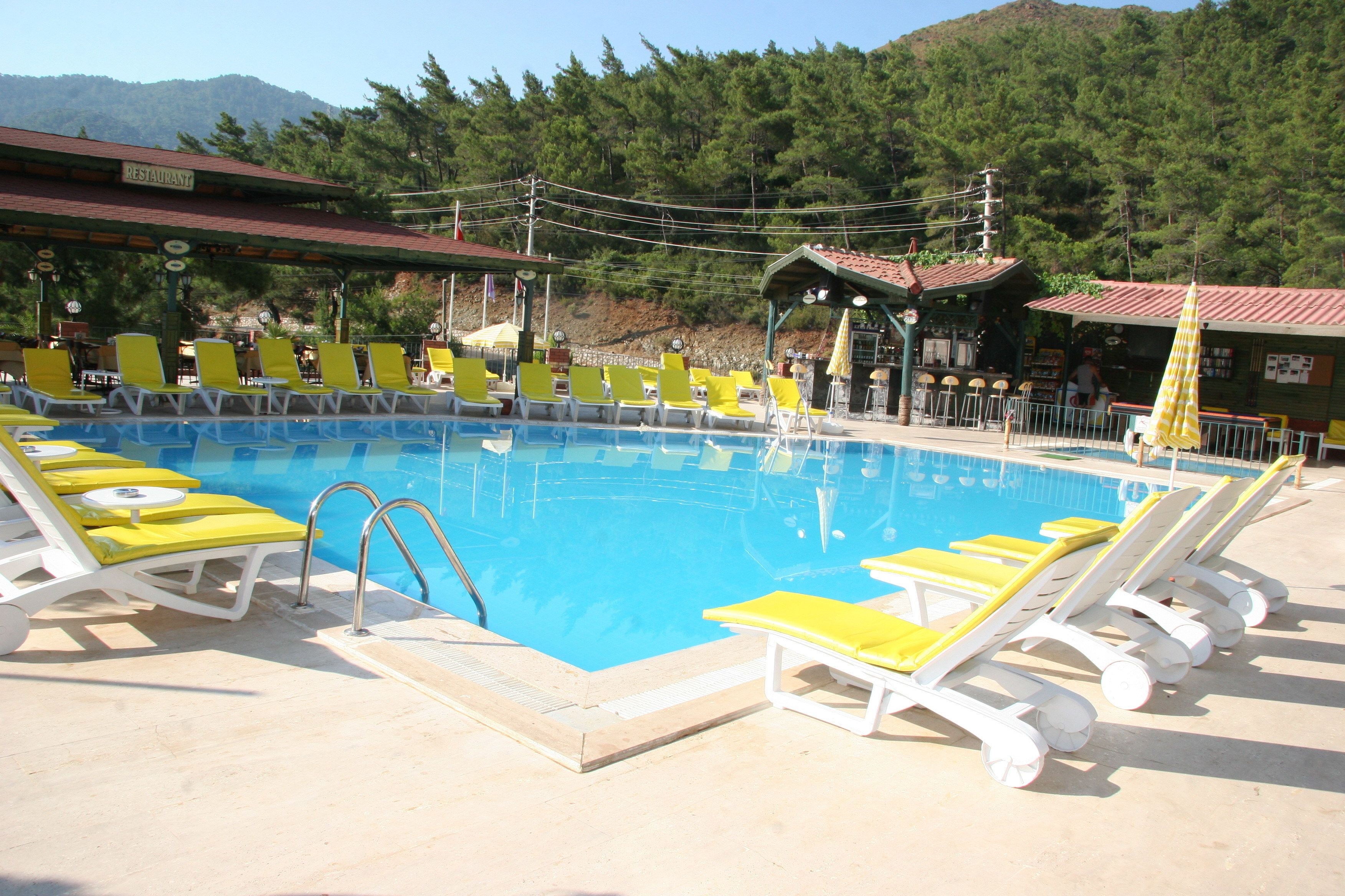 Piscina com cadeiras de sol amarelas e brancas, um restaurante e montanhas ao fundo.