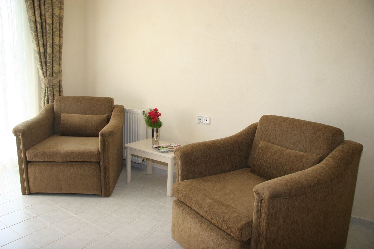 A room with two brown armchairs, a small white table with a vase of flowers, and a radiator.