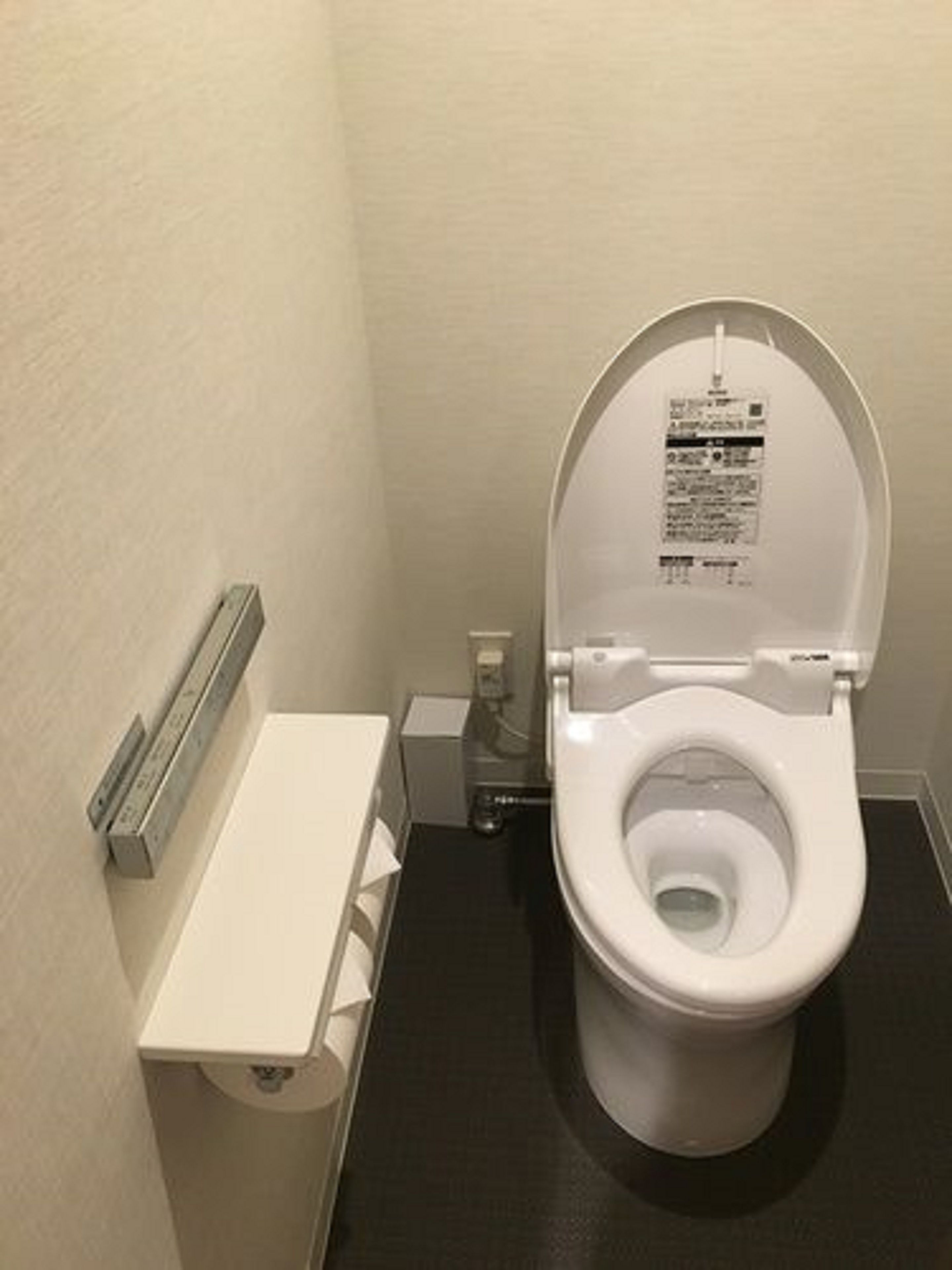 Un inodoro estándar con asiento y tapa blancos, un secador de manos instalado en la pared y un pequeño cubo de basura.