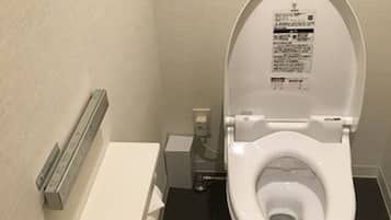 Un inodoro estándar con asiento y tapa blancos, un secador de manos instalado en la pared y un pequeño cubo de basura.