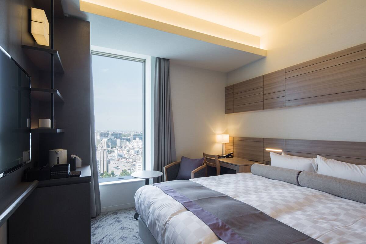 A modern hotel room with a large bed, a desk, a chair, a TV, and a view of the city.