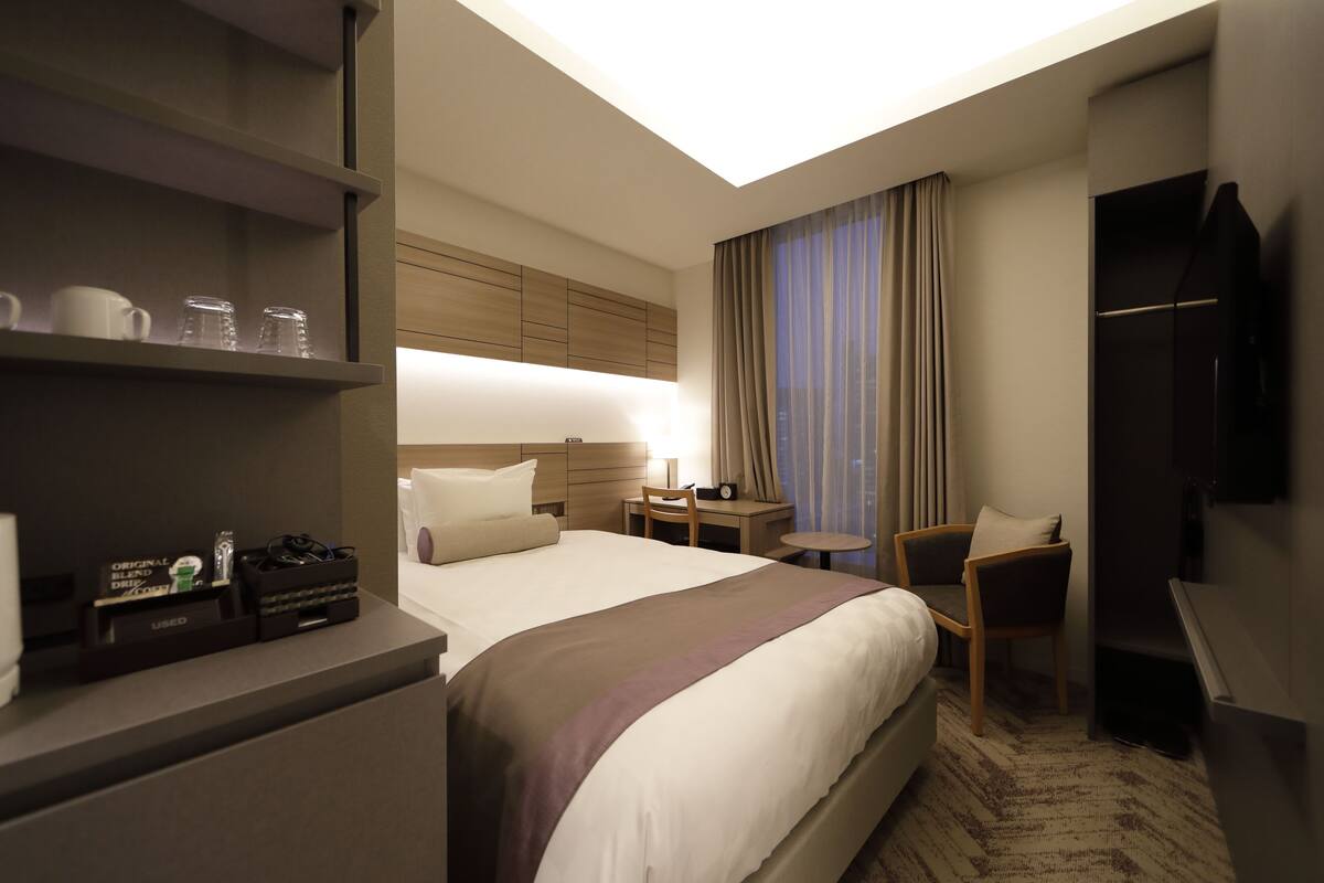 A hotel room with a large bed, a desk, a chair, a TV, and a shelf with items.