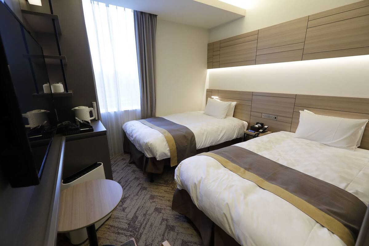 A hotel room with two beds, a desk, a chair, a TV, and a window with curtains.