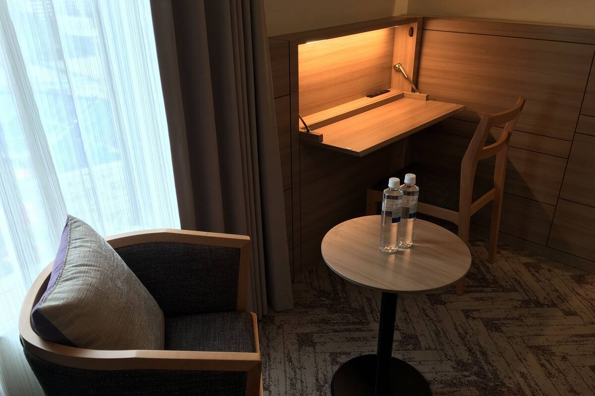 A hotel room with a corner desk, a round table with two water bottles, a cushioned chair, and a view of the city through large windows.