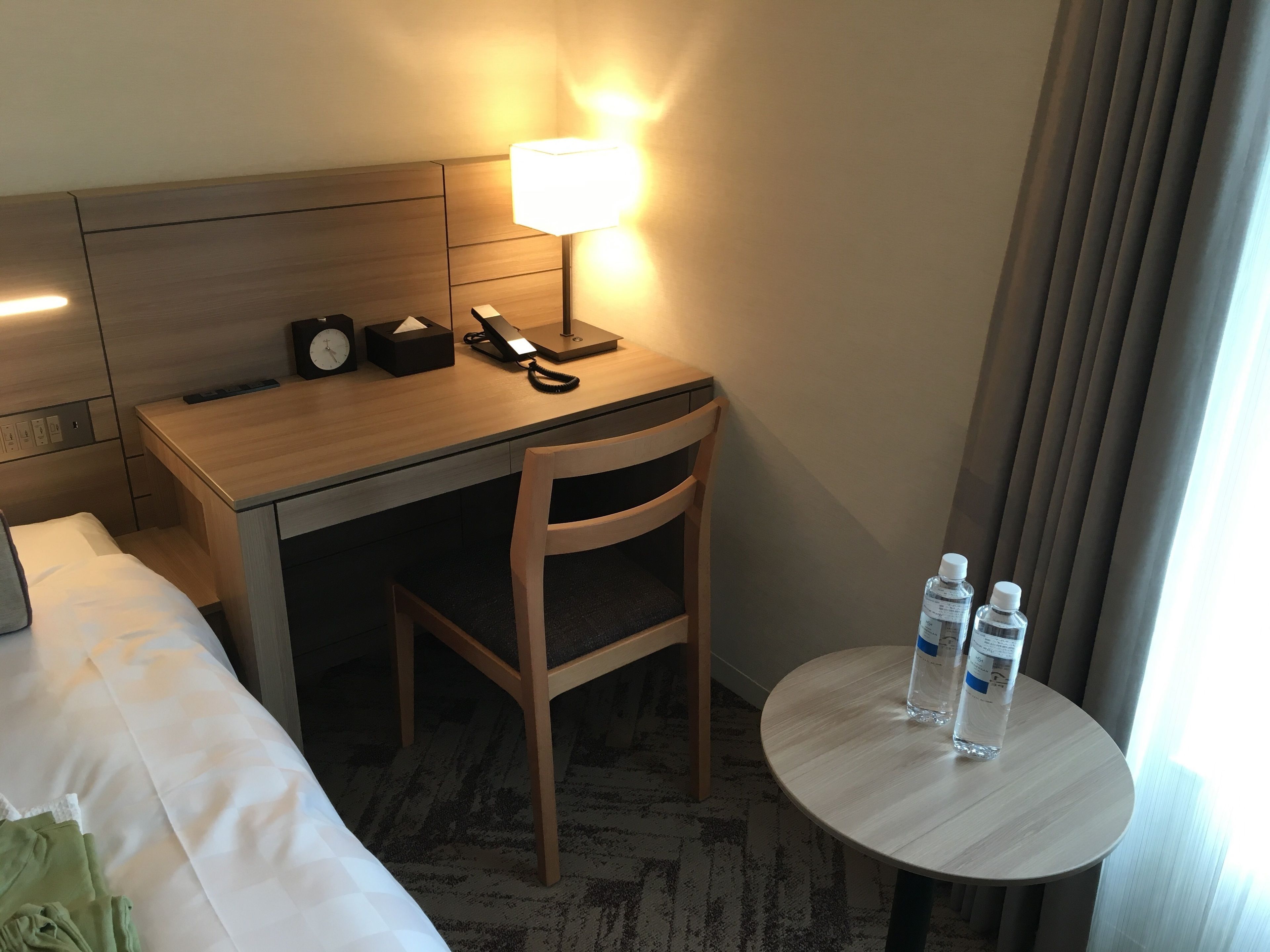 A hotel room with a bed, a desk with a lamp, a chair, and a small table with water bottles.