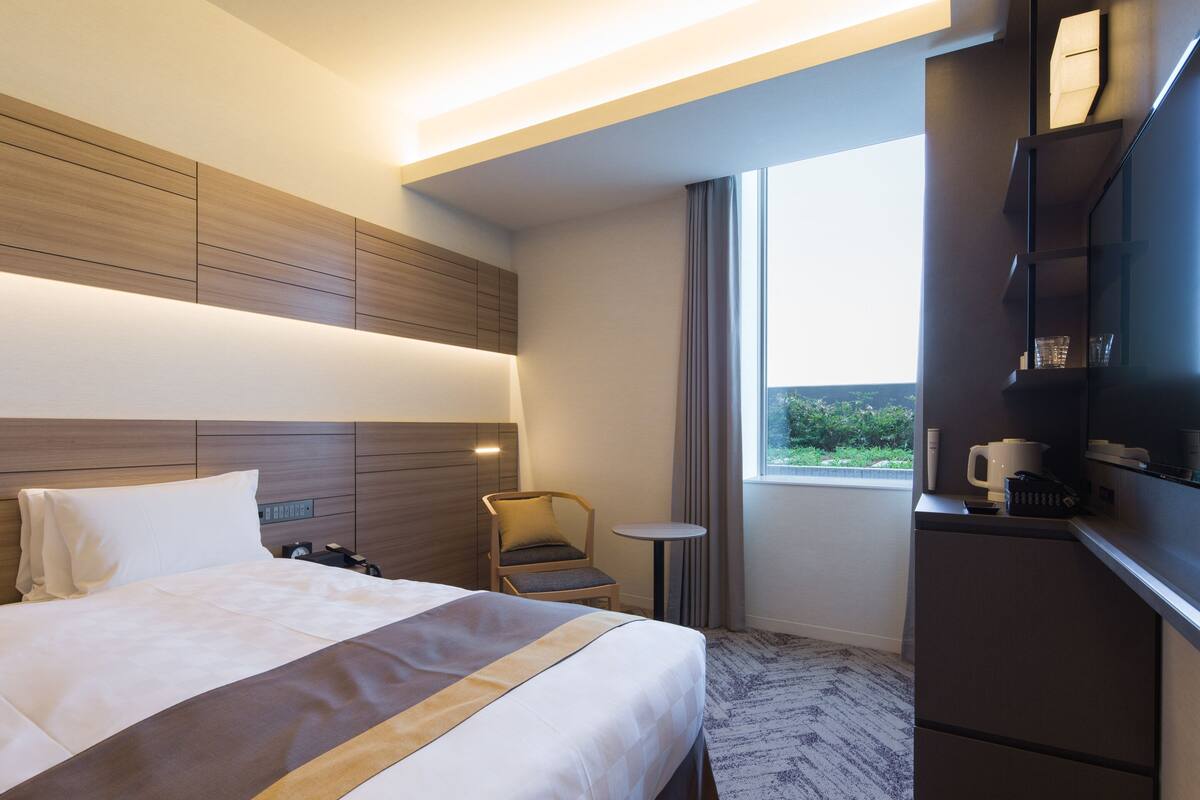 A modern hotel room with a large bed, a small table, a chair, and a built-in desk with a mirror.