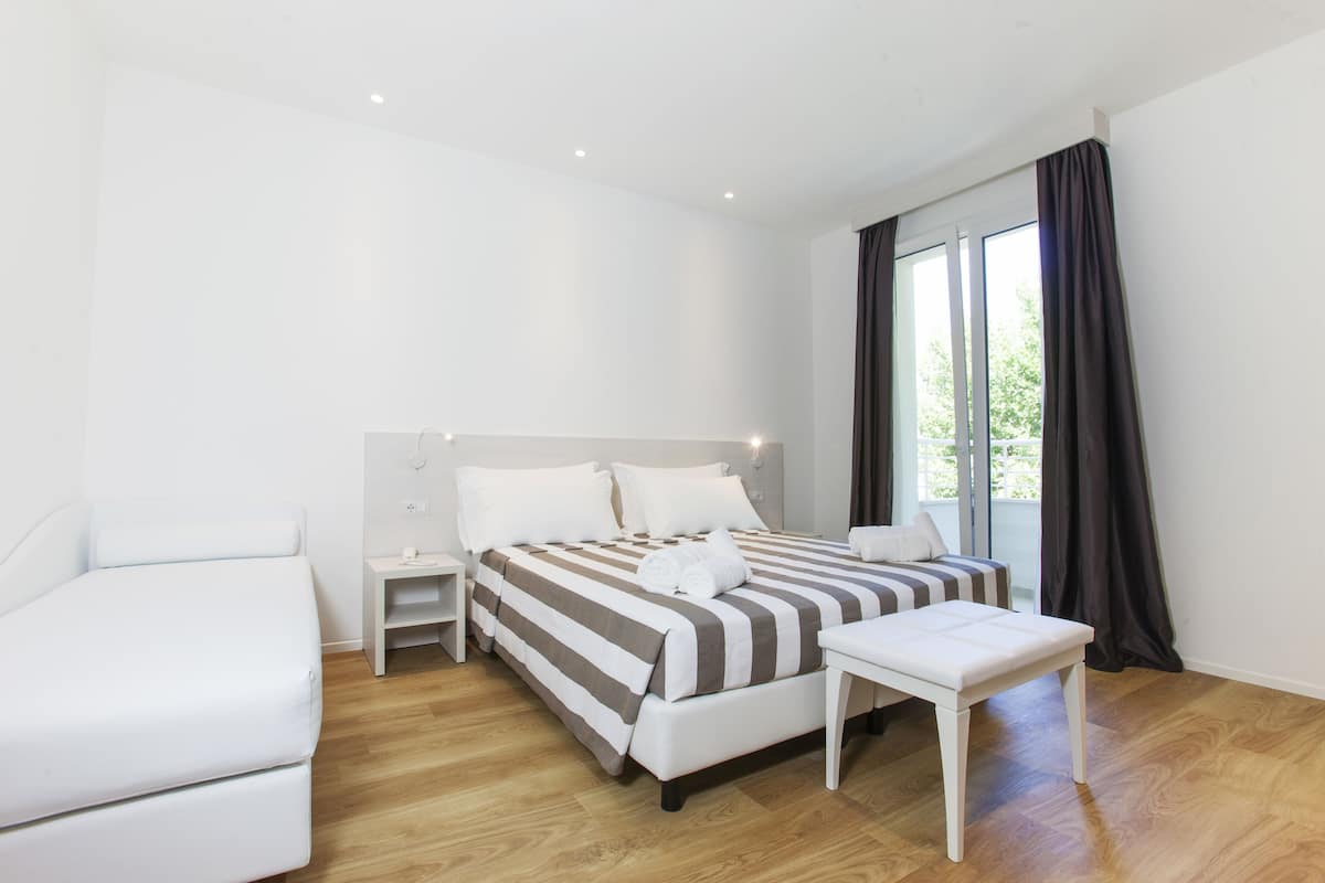 A modern hotel room with a large bed, a bedside table, a small desk, and a bench.