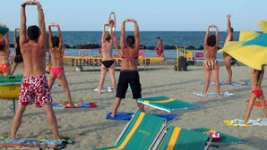 En grupp människor utför en stretchövning på en sandstrand, med strandstolar och parasoller i bakgrunden.