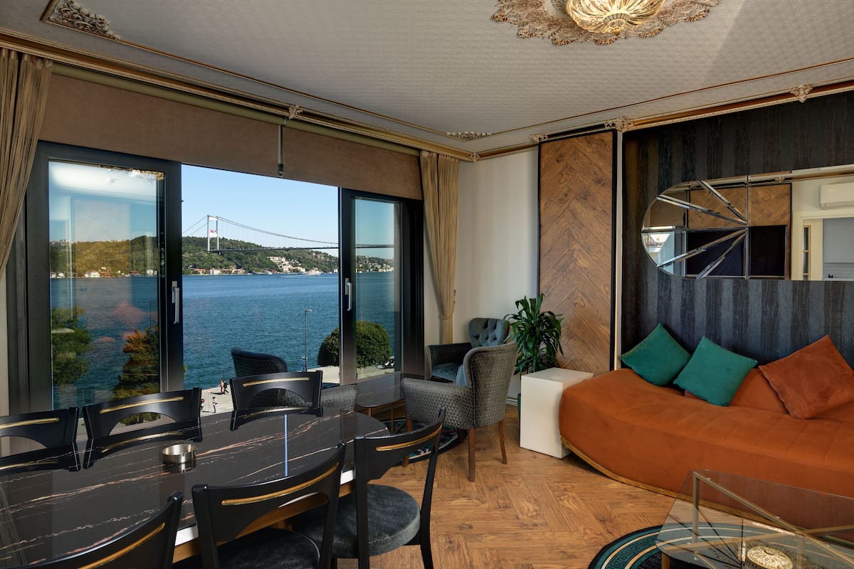 A modern living room with a large window overlooking a body of water and a bridge, furnished with a dining table, chairs, a sofa, and a coffee table.