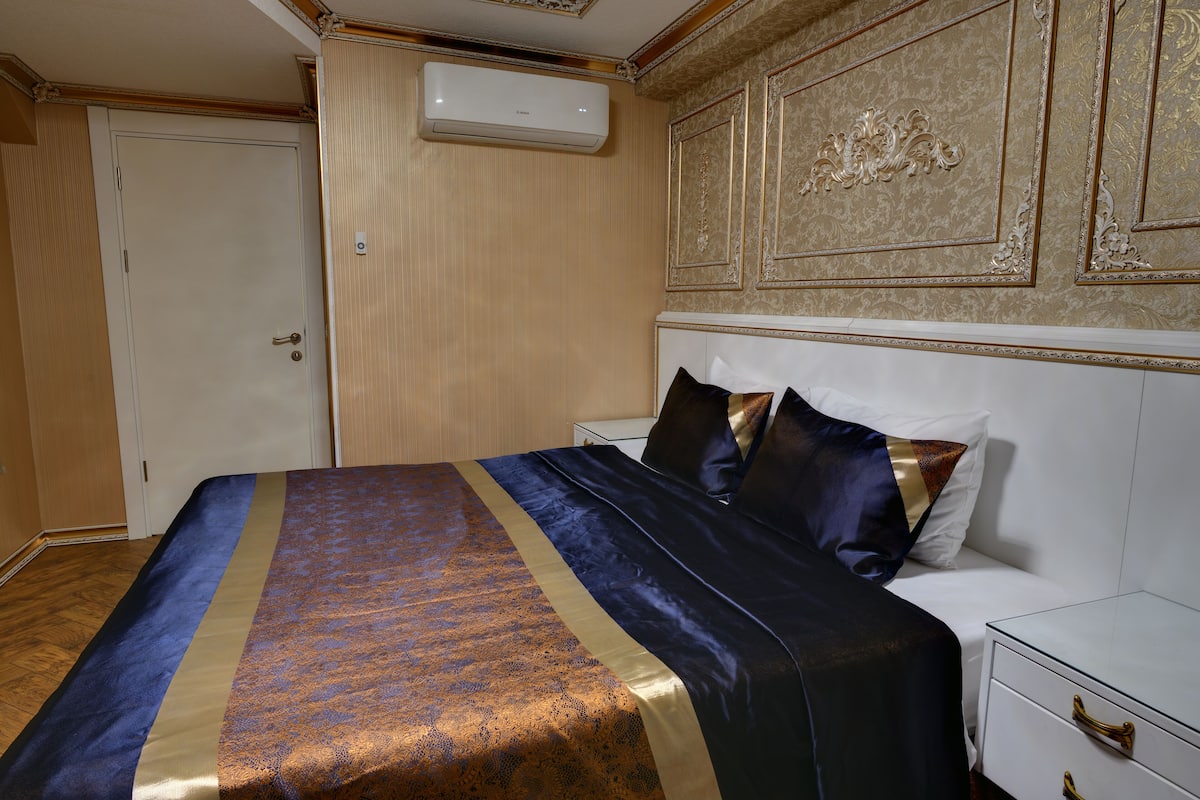 A neatly made bed with a patterned bedspread, a bedside table with drawers, and a wall-mounted air conditioner.