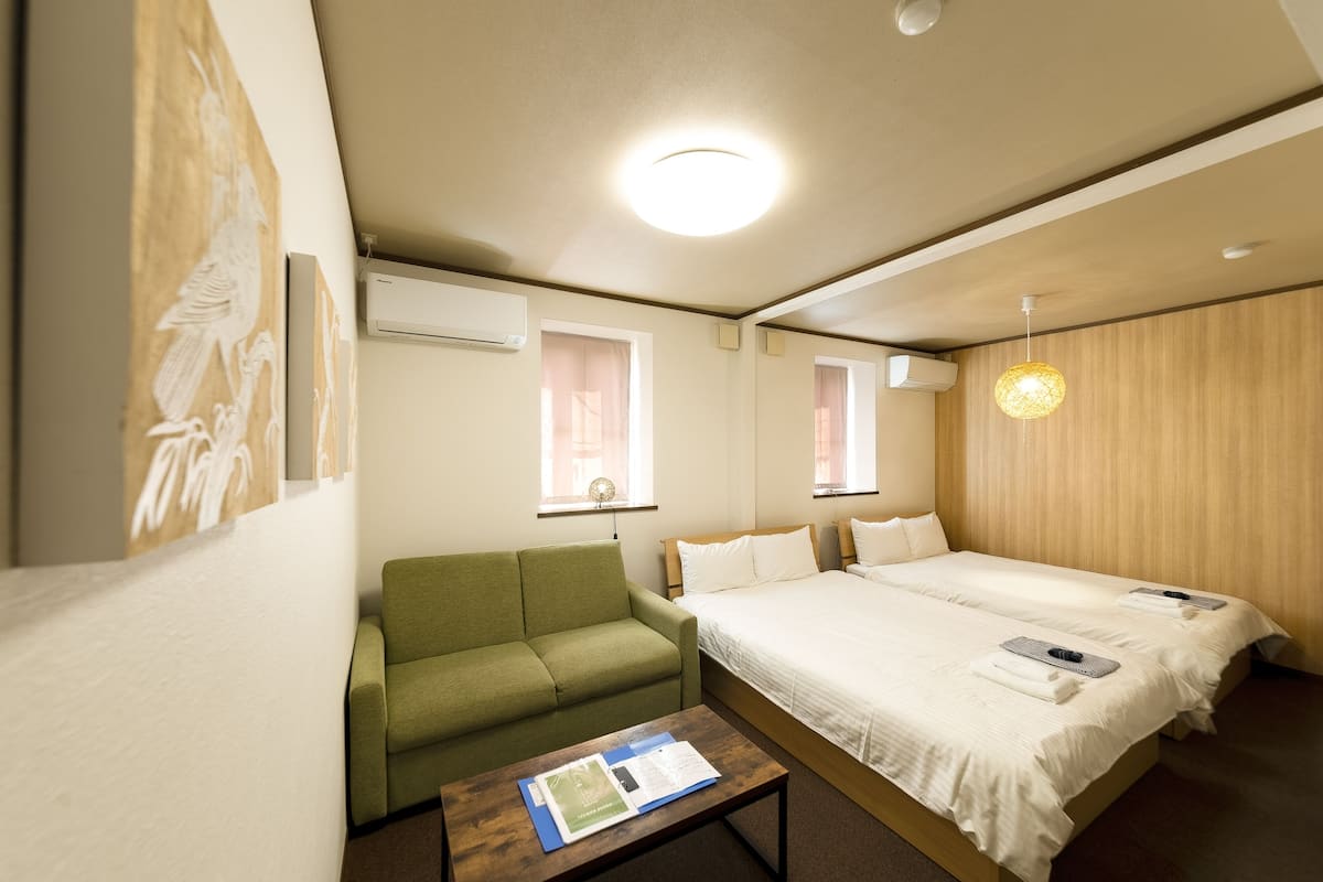Quarto de hotel com uma cama de solteiro, um sofá verde, cabeceira de madeira e uma luminária no teto.