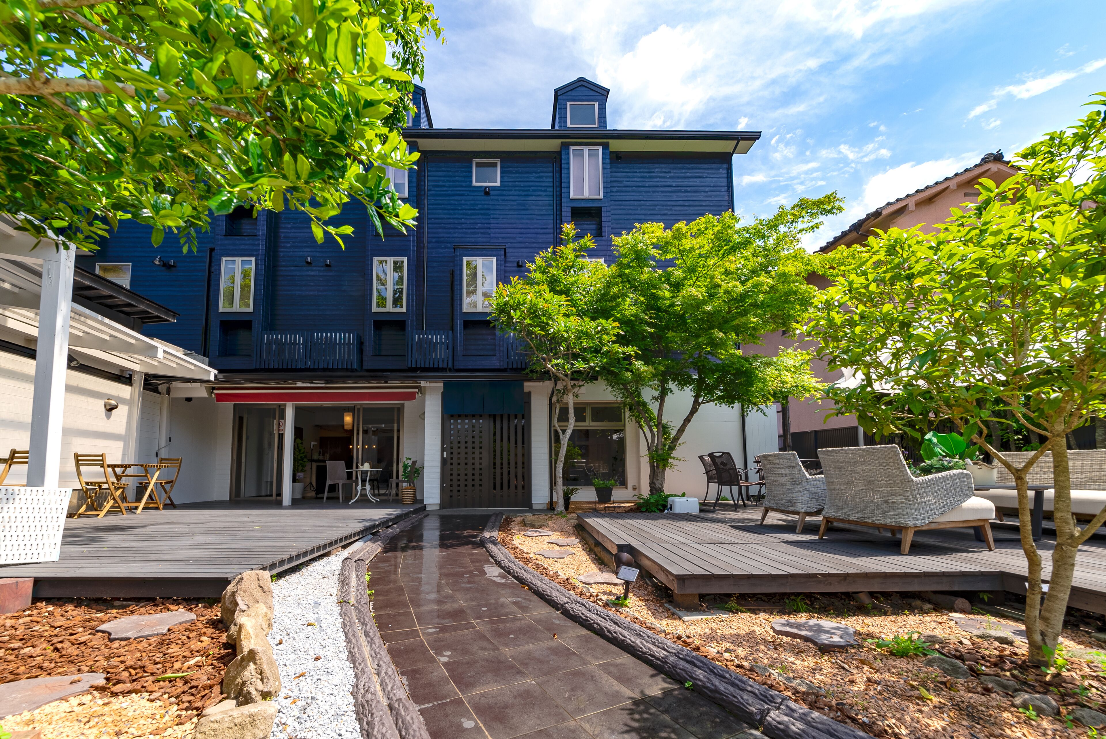 A two-story building with a dark blue exterior, a covered patio area with wicker furniture, and a small garden with a tree and shrubs.