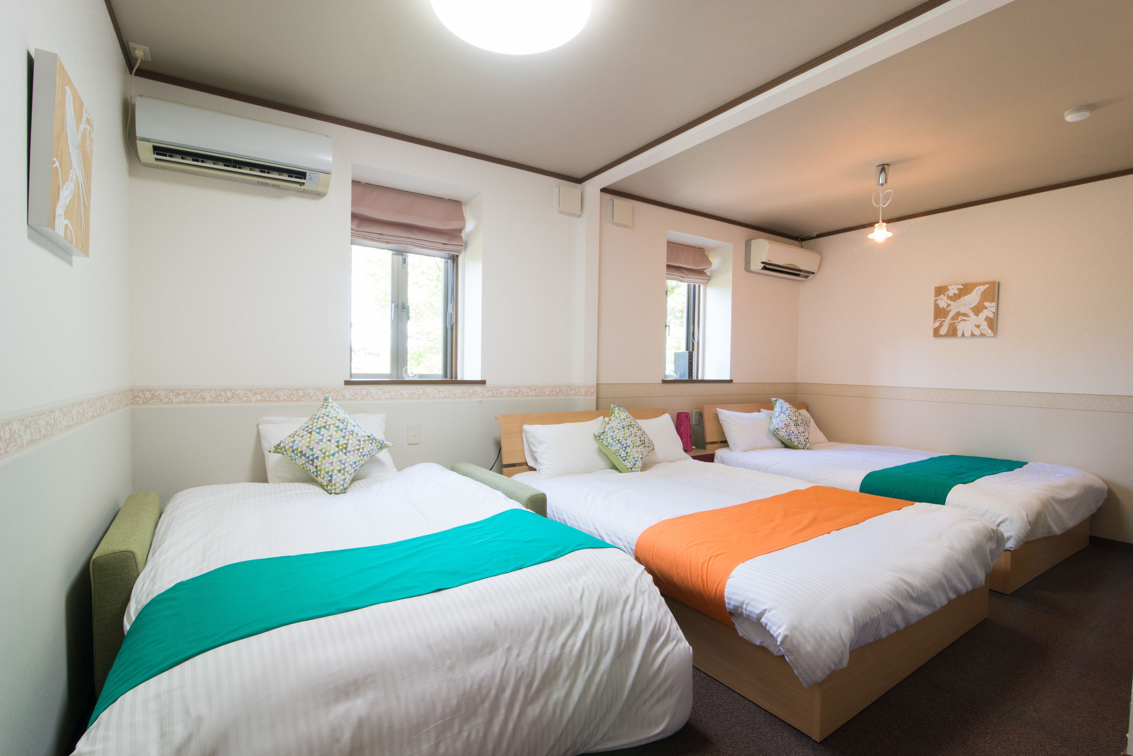 A hotel room with three beds, each with a different colored blanket, air conditioning units, and a window with a blind.