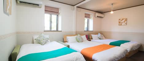 A hotel room with three beds, each with a different colored blanket, air conditioning units, and a window with a blind.