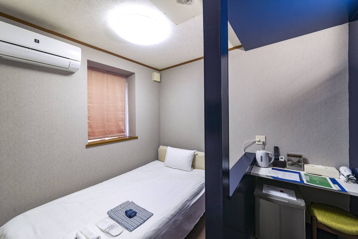 Uma cama de solteiro com edredom branco, uma mesa de cabeceira com luminária, uma escrivaninha com computador e uma cadeira, e uma janela com cortina.