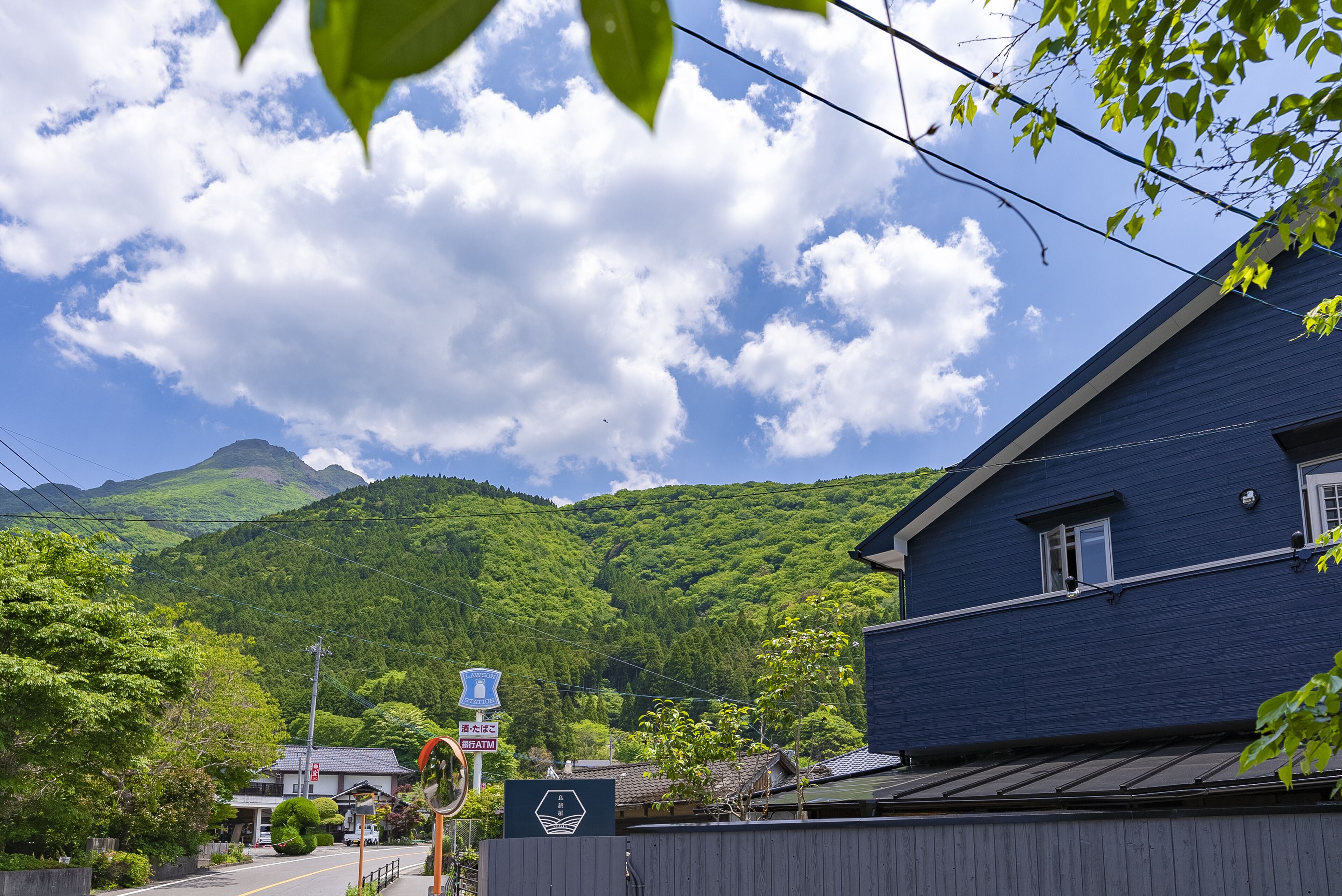 A residential area with a blue house, a small building with a sign, and lush green hills in the background.