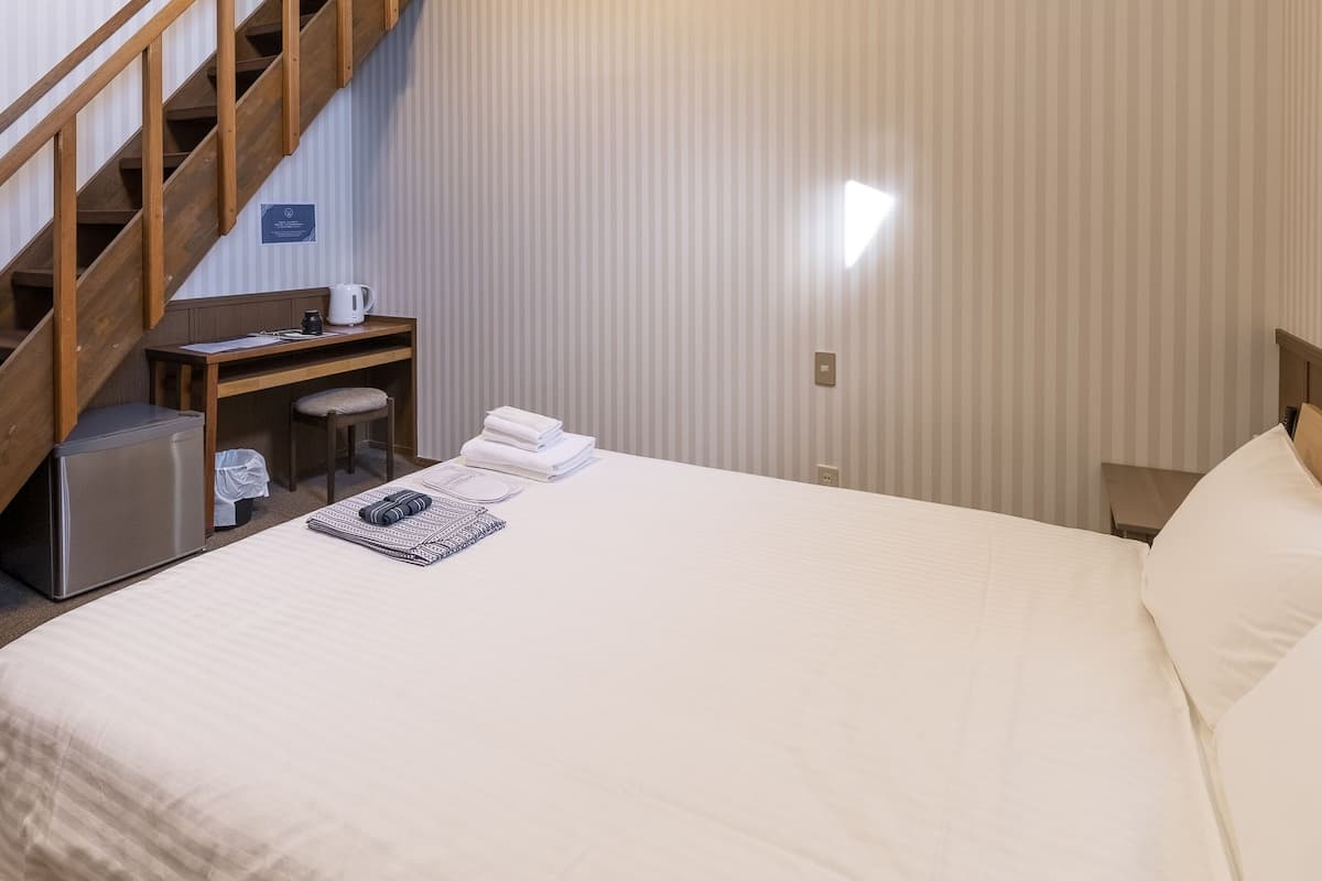 Quarto de hotel com cama, escrivaninha, cadeira, cafeteira e escada de madeira.