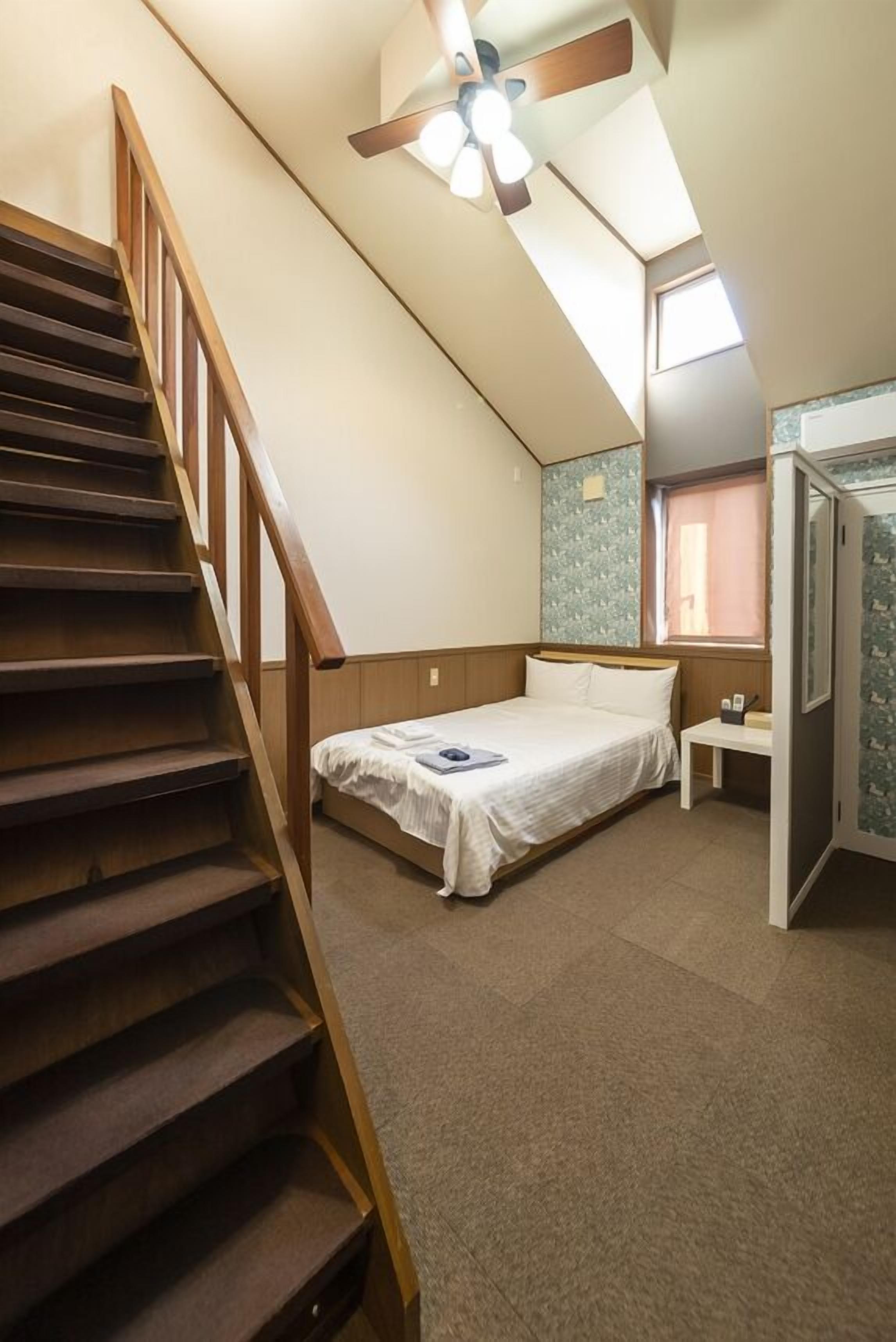 A bedroom with a bed, a nightstand, a wooden staircase, and a ceiling fan.