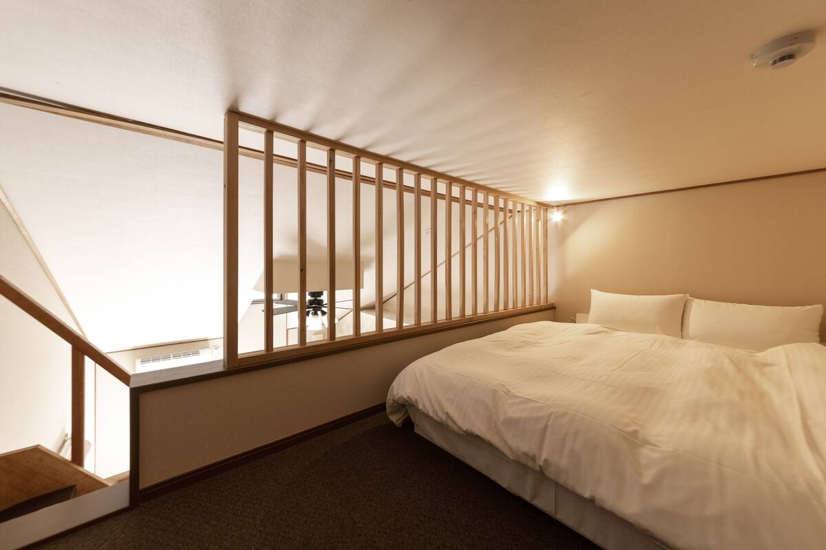 Quarto com cama, janela emoldurada em madeira e escada que leva a um andar superior.