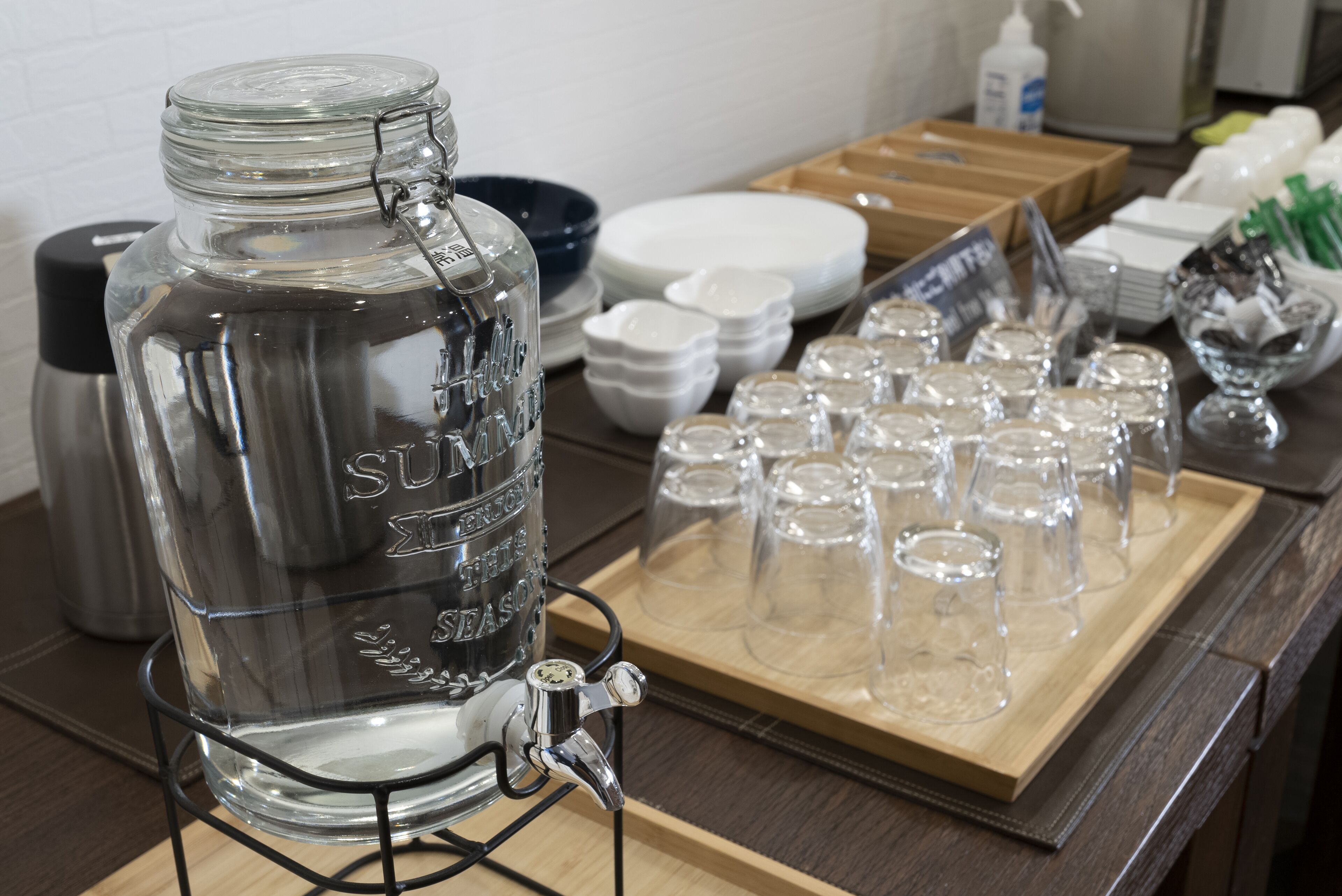 Een waterdispenser met een metalen kraan, omringd door glazen bekers en een schaal lege glaasjes.