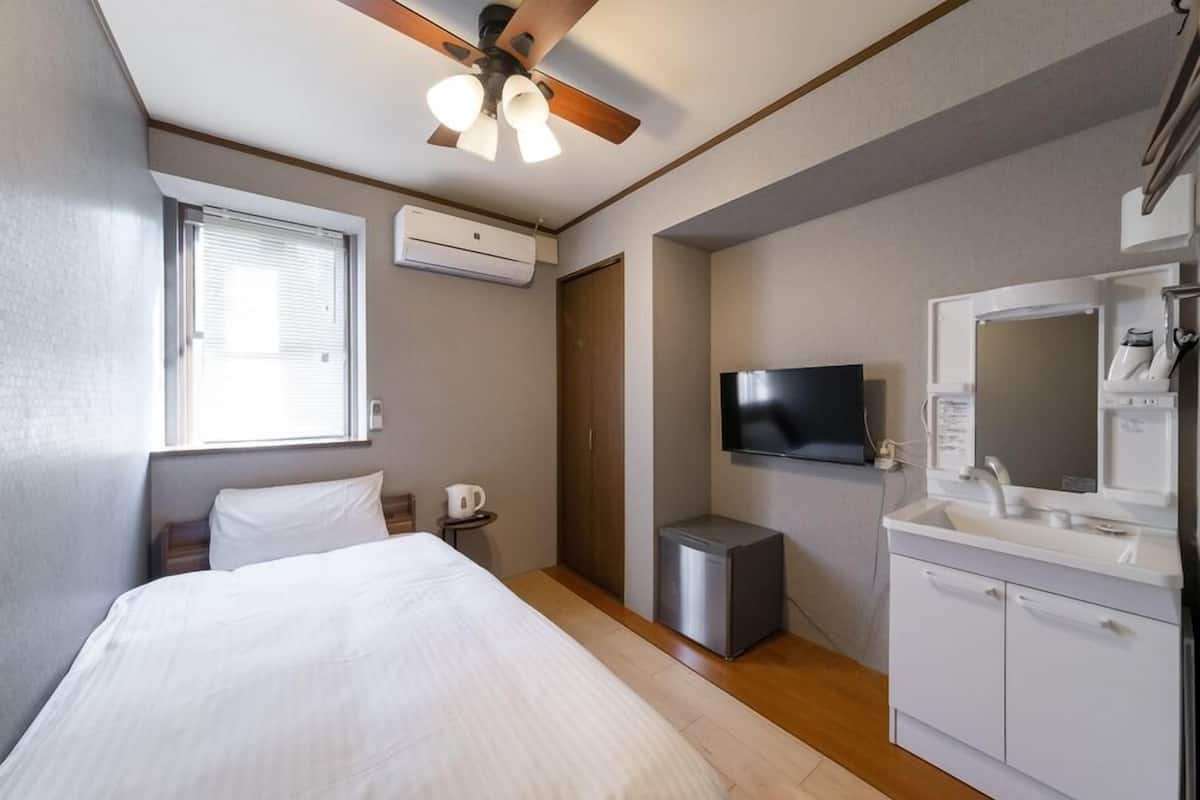 Quarto com cama, ventilador de teto, televisão, mesinha de cabeceira, criado-mudo, janela com persianas e banheiro com pia e espelho.