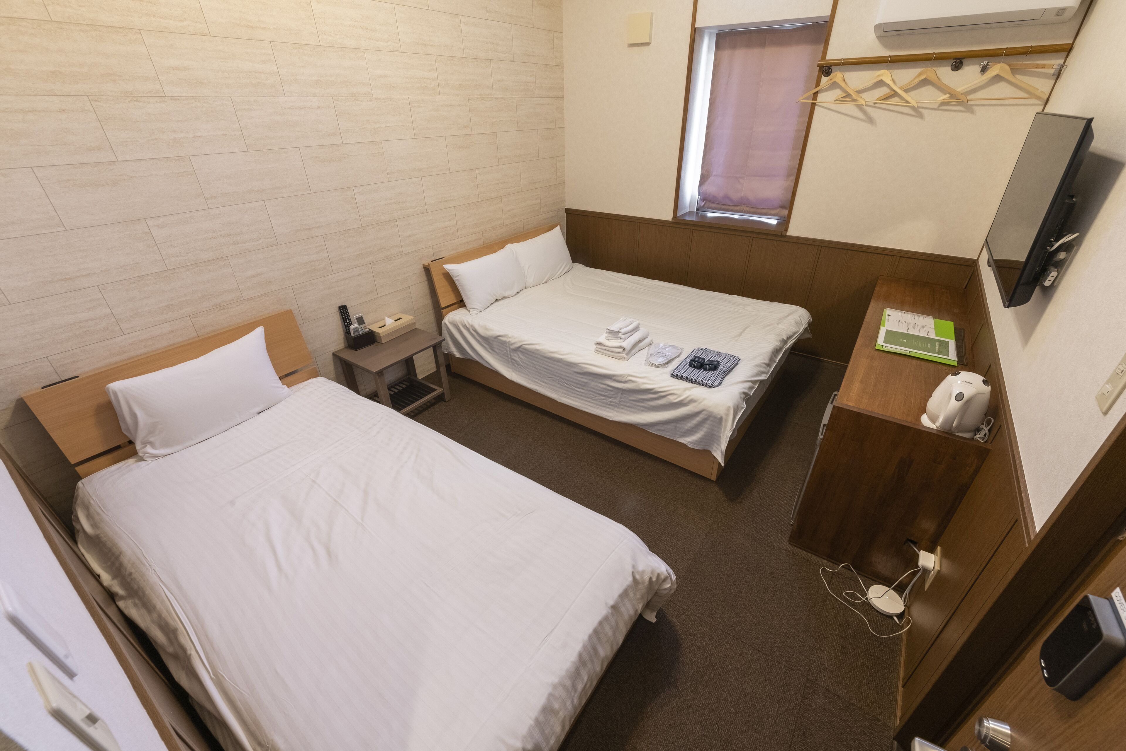A hotel room with two beds, a wooden headboard, a small bedside table with a phone and a lamp, a wooden cabinet with a mirror and a clock, and a window with a curtain.