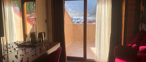 A room with a wooden desk, a chair, a red sofa, and a large sliding glass door leading to a balcony with a view of a lake and mountains.