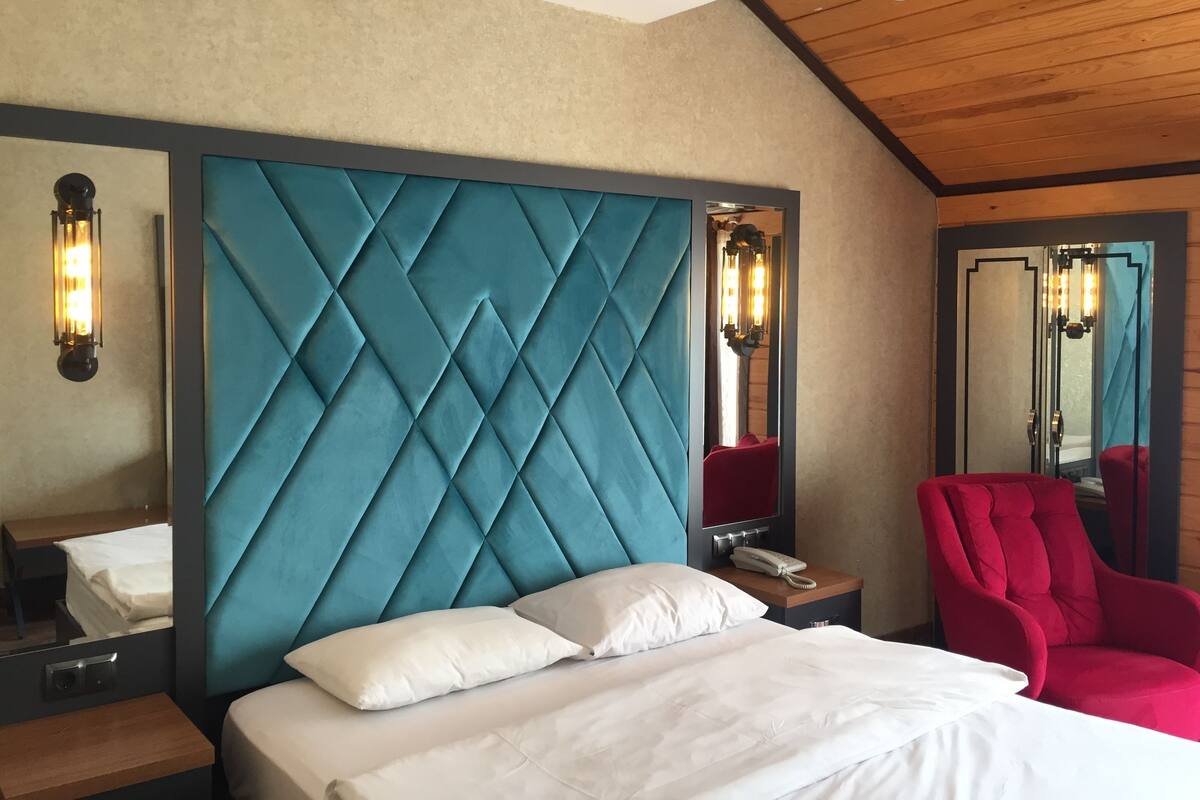 A hotel room with a large bed, a wooden ceiling, a blue headboard, a red armchair, and a nightstand with a phone.