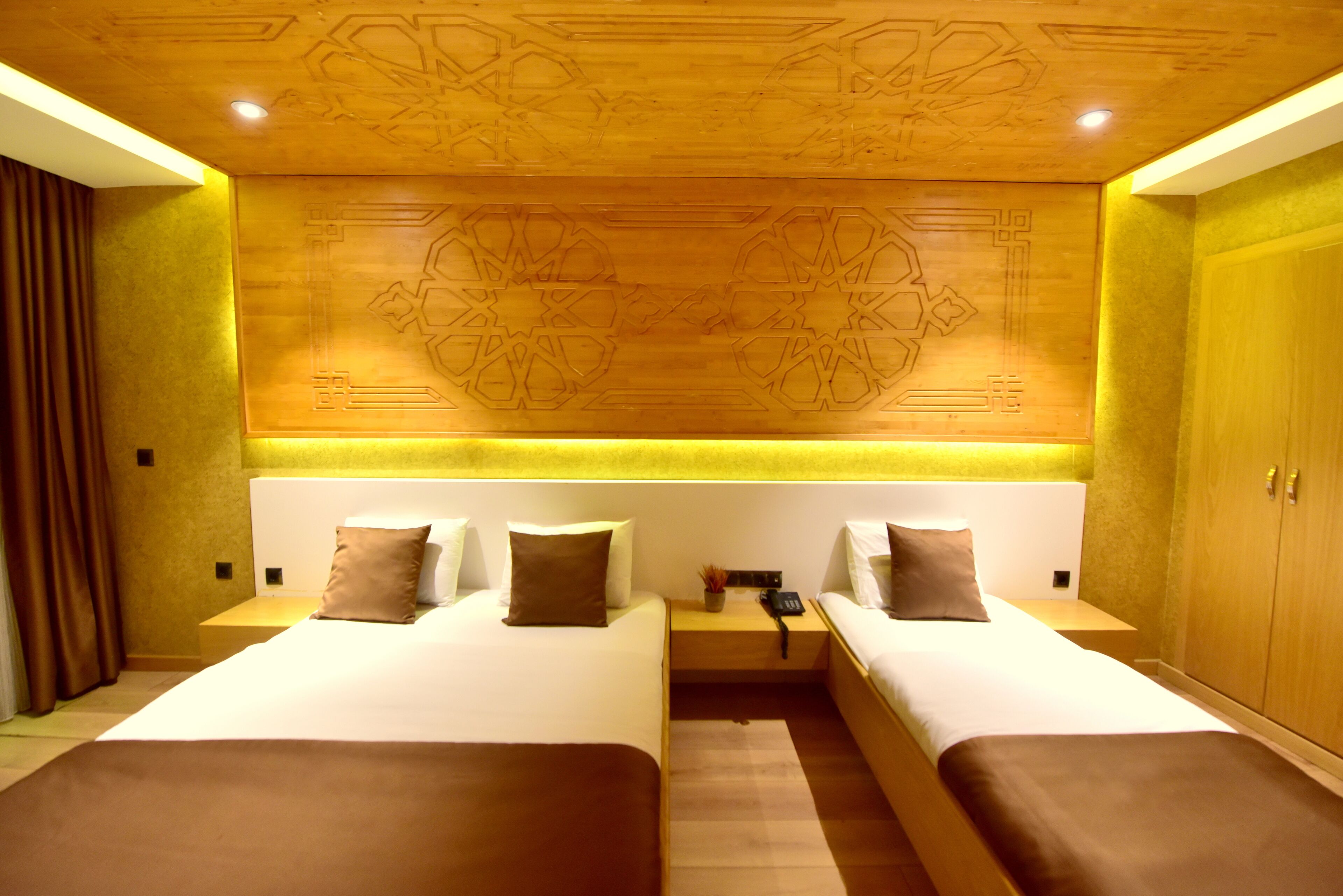 A hotel room with two beds, a wooden headboard with geometric patterns, a bedside table with a lamp, and a wardrobe.