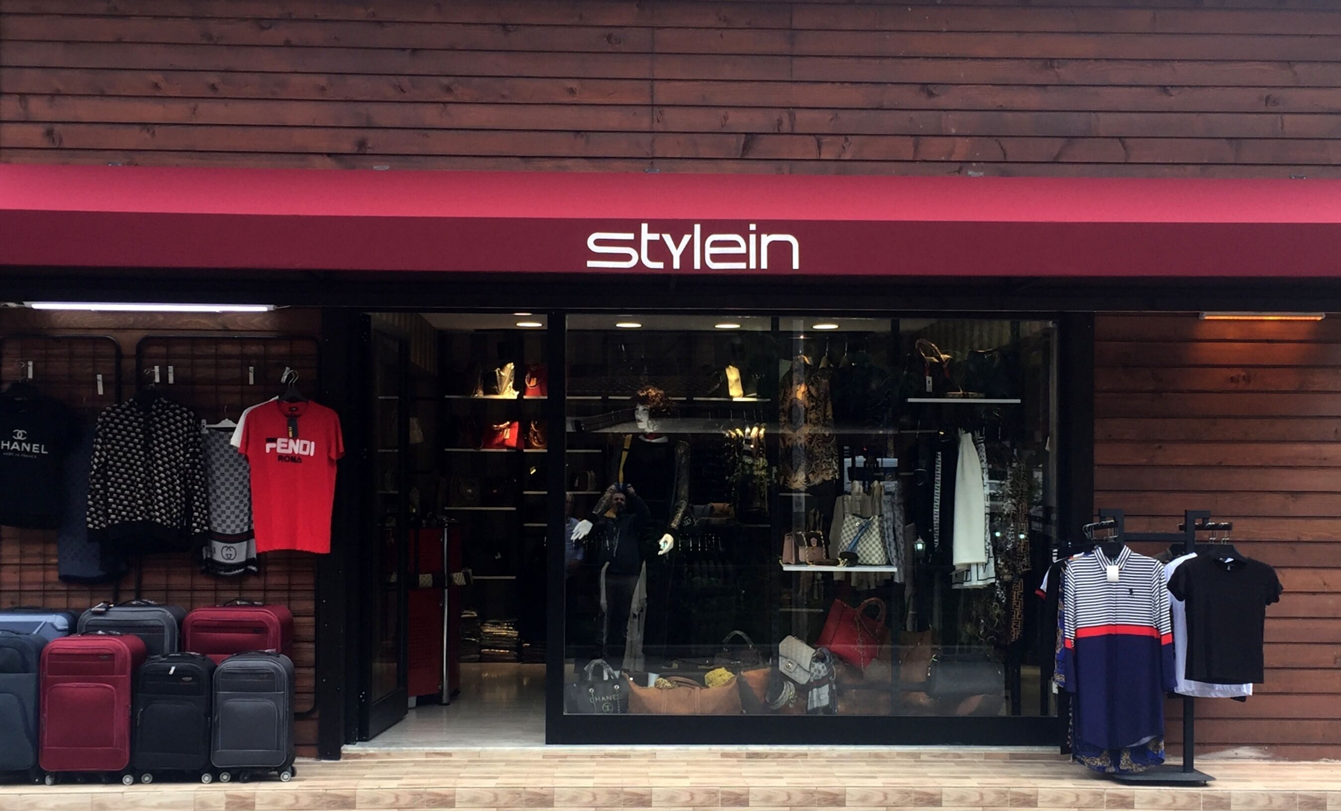 Un escaparate con toldo rojo y la palabra “Stylein” sobre la entrada. En el interior, se exhiben diferentes prendas de vestir y bolsos en la ventana.