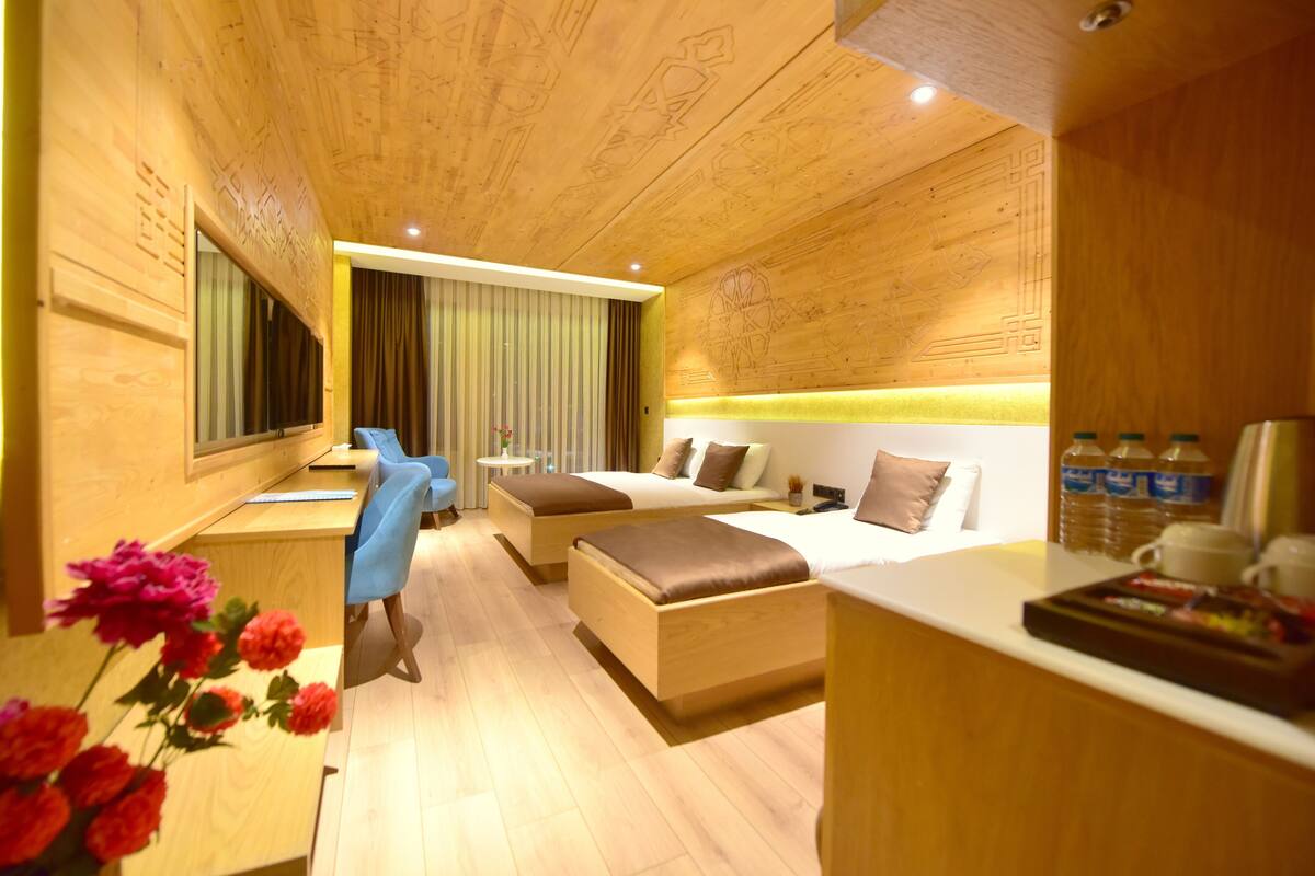 A modern hotel room with a wooden ceiling, a desk with a chair, a bed with a headboard, and a bedside table.