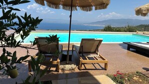 Zona junto a la piscina con dos sillones de descanso y un paraguas de paja, con vista a una piscina azul cristalina y montañas distantes.