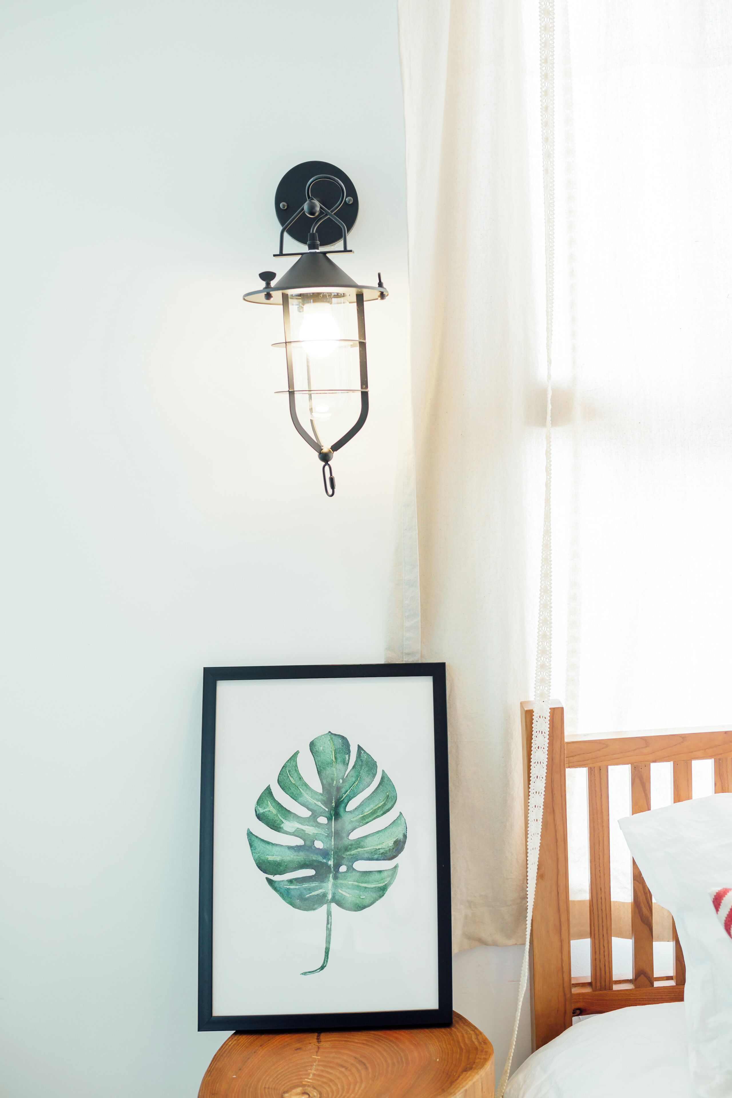 Une œuvre d’art représentant une feuille, encadrée, posée sur une table de chevet en bois, un luminaire fixé au mur et un berceau en bois avec un coussin blanc.