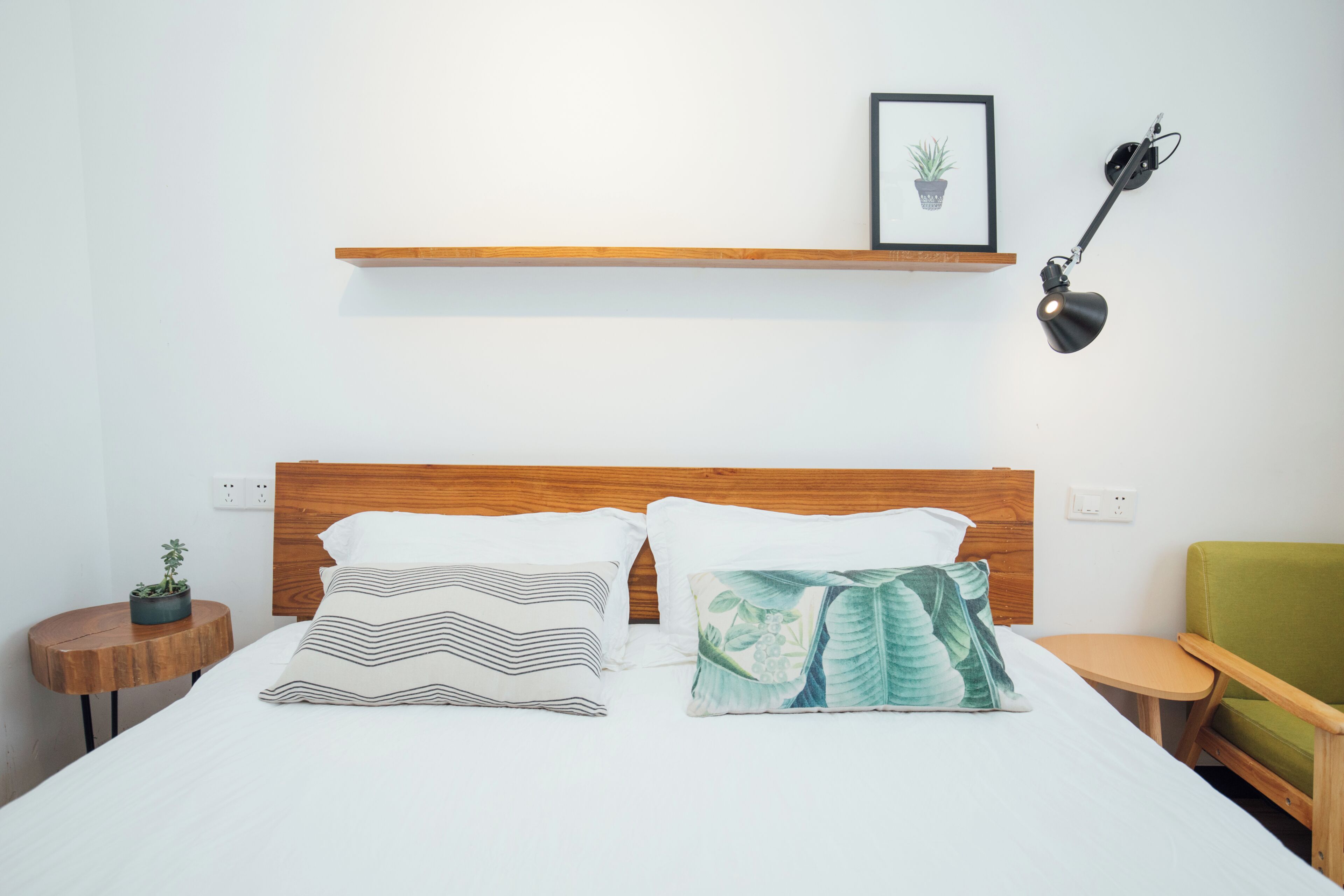 Una cama bien hecha con dos almohadas, una mesita de noche con una planta, una pequeña mesa redonda, una silla verde, un estante con un cuadro enmarcado y una lámpara de escritorio.