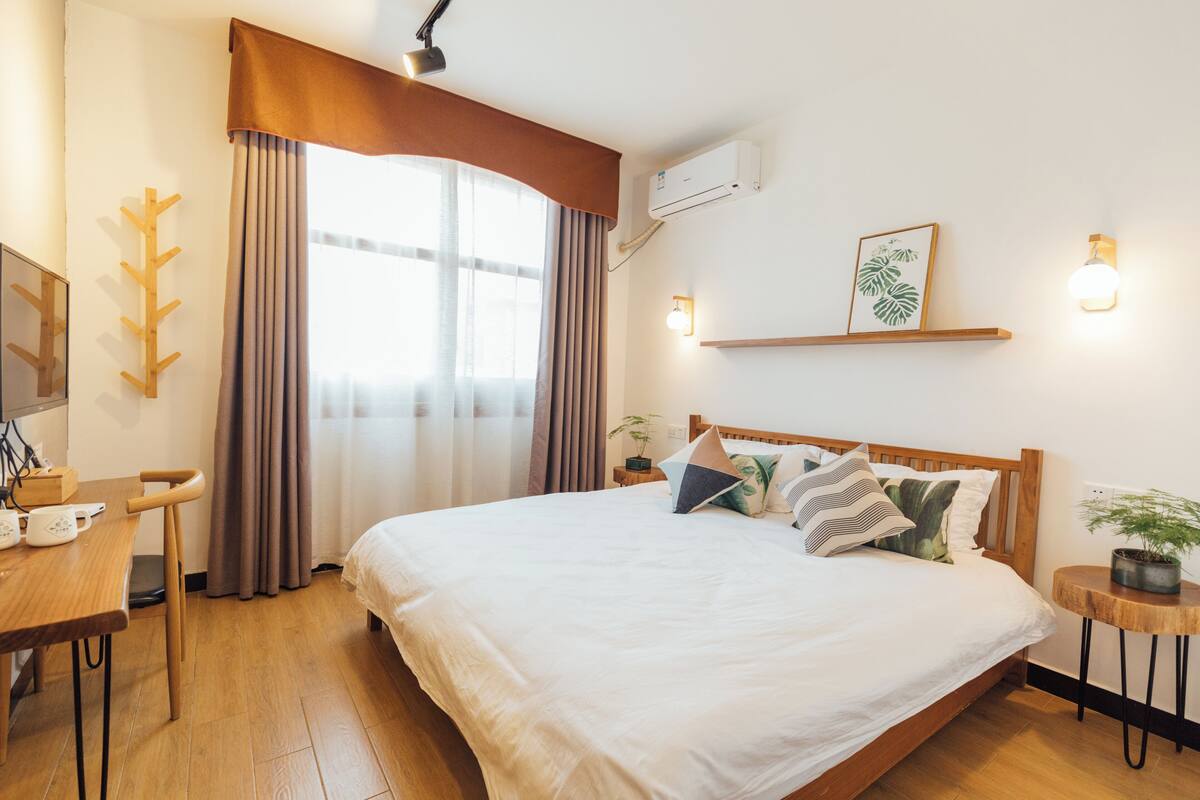 A hotel room with a bed, bedside table, lamp, air conditioning unit, and a window with curtains.