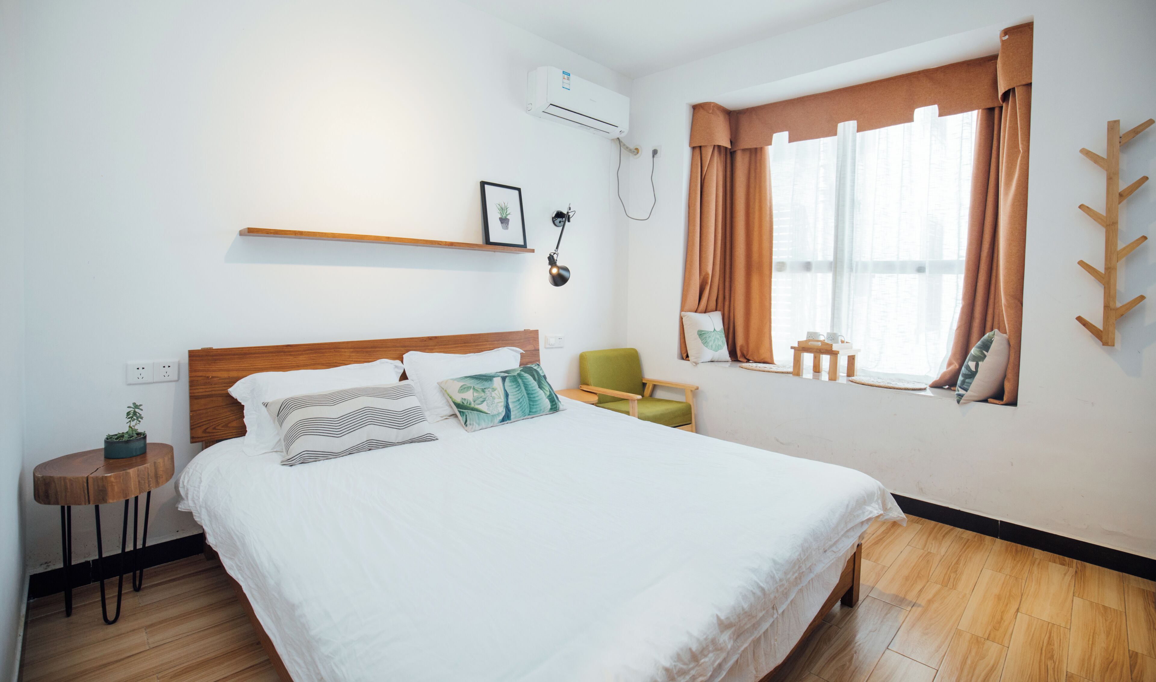 A neatly arranged hotel room with a bed, a small table, a chair, a shelf, and a window with curtains.