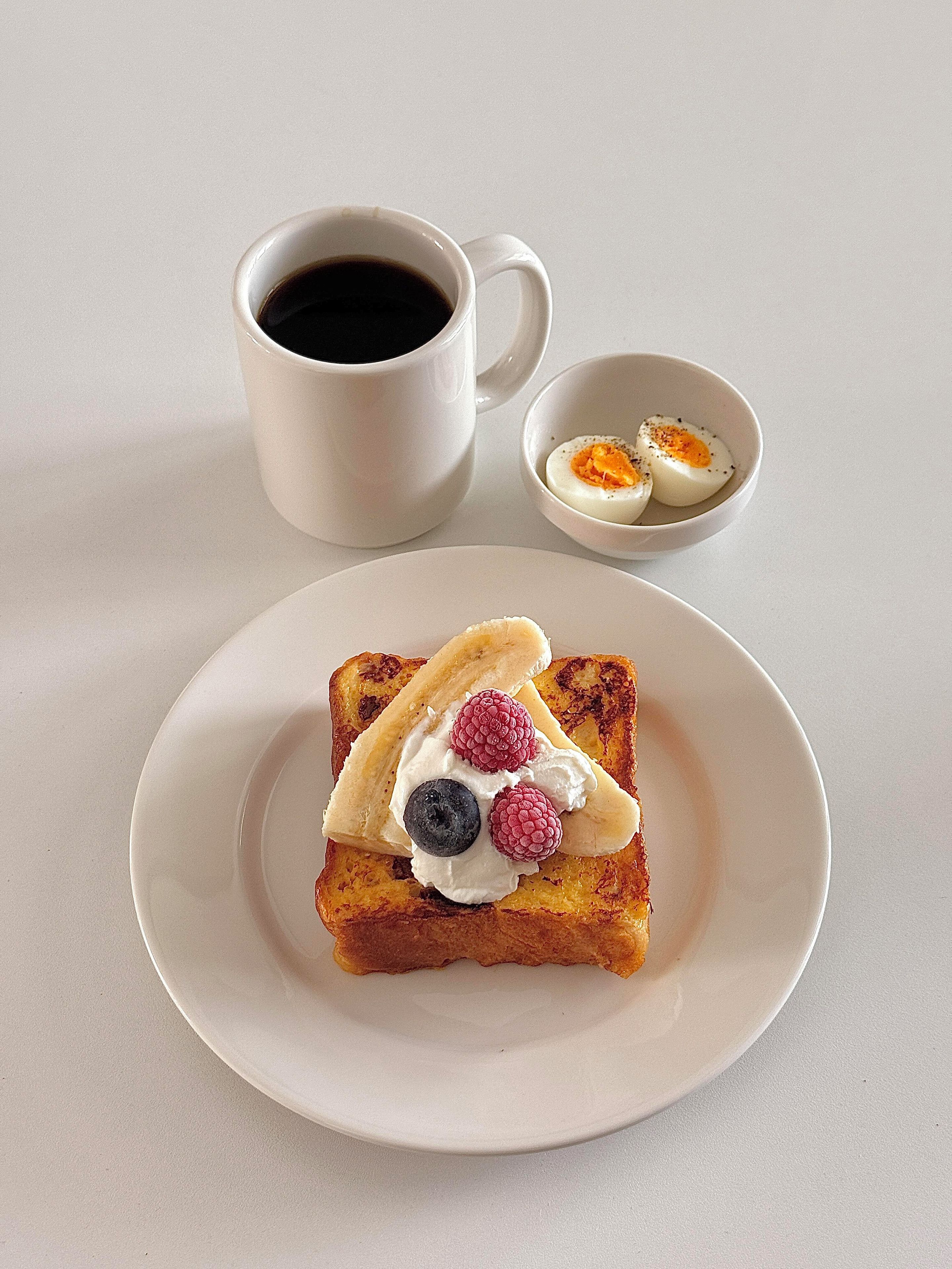 Prato com torradas cobertas de chantilly, framboesas e um mirtilo, acompanhado de uma xícara de café preto e uma tigela pequena de ovos cozidos fatiados.