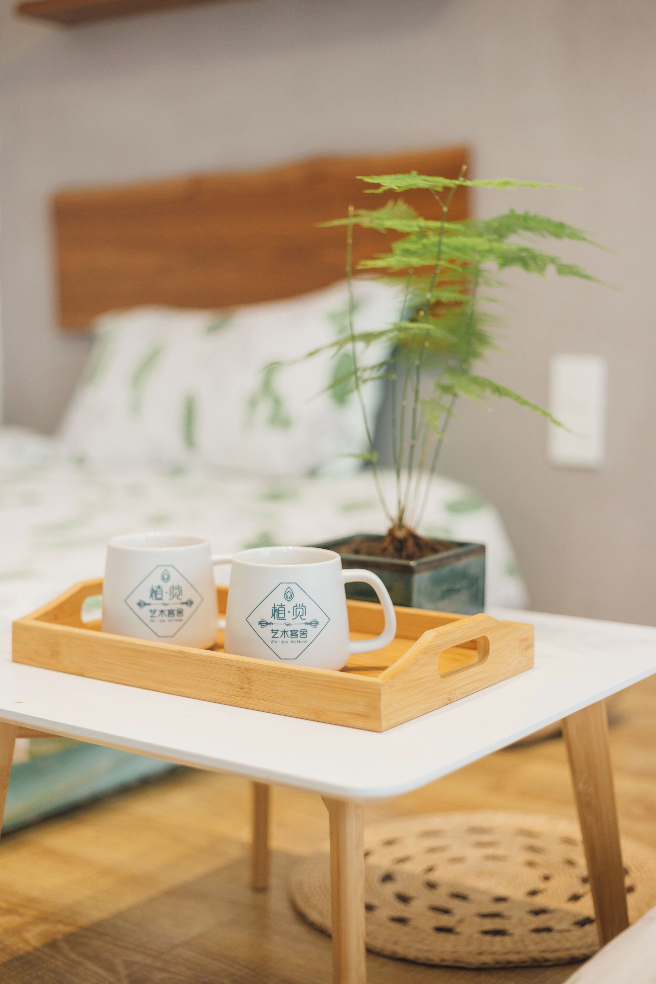 A bed with a wooden headboard, a potted plant, and a tray with two white mugs on a white table.